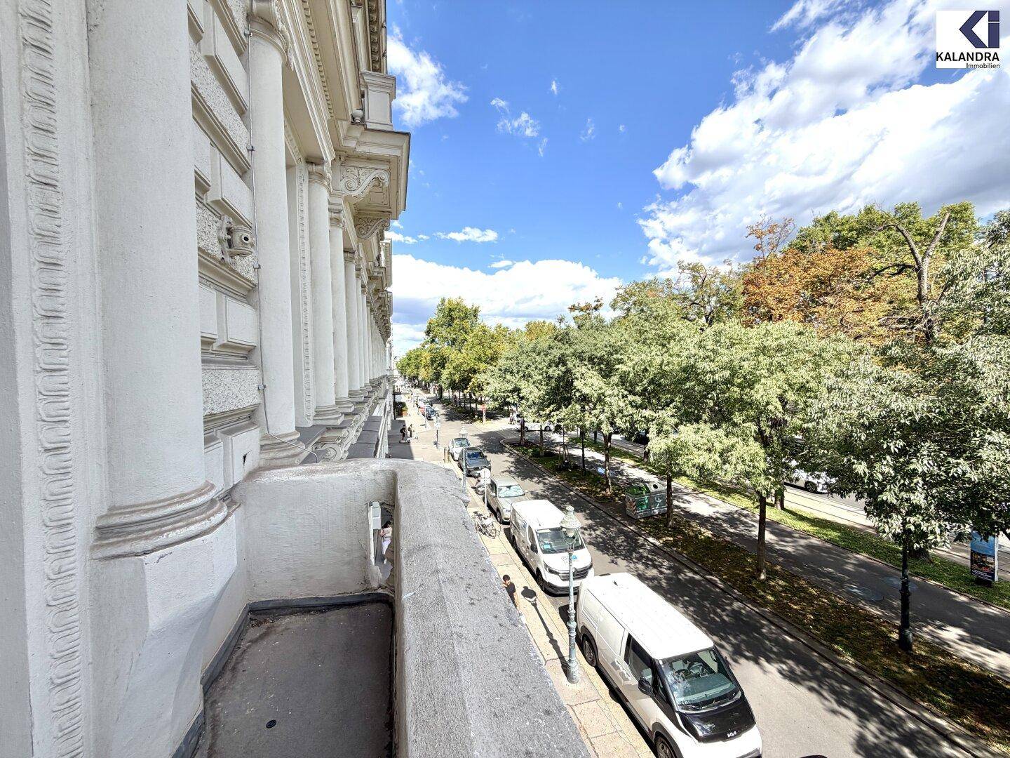 Schmaler Balkon mit Blick auf eine von Bäumen gesäumte Straße und historische Gebäude unter blauem Himmel.