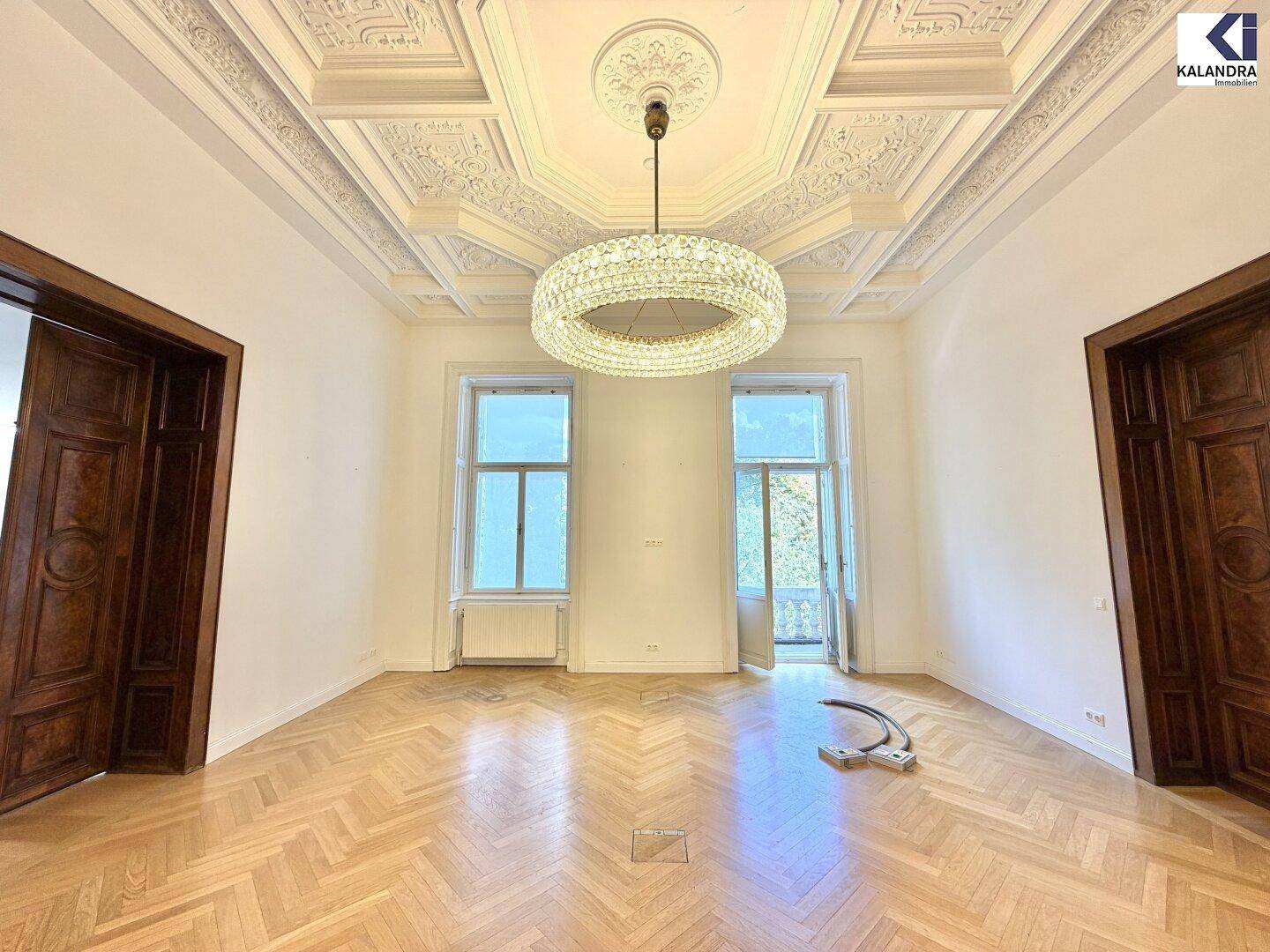 Heller Raum mit Fischgrätparkett, hohen Decken und einem modernen Kronleuchter, mit Zugang zu einem Balkon.