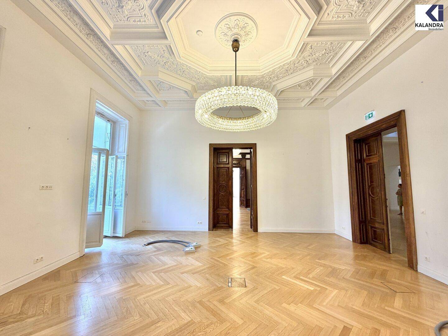 Geräumiger Raum mit Fischgrätparkett, hohen Decken und einem modernen Kronleuchter, mit mehreren Holztüren.