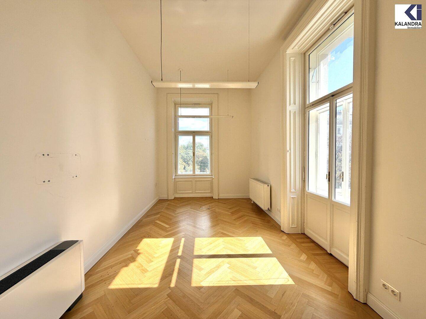 Heller, leerer Raum mit Fischgrätparkett und hohen Fenstern, die viel Tageslicht hereinlassen.