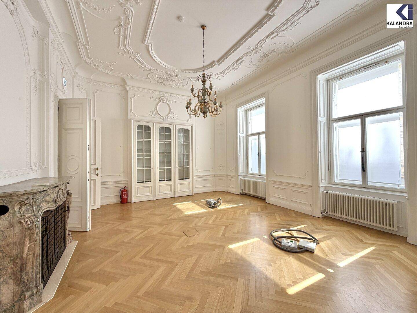 Eleganter Raum mit Fischgrätparkett, hohen Stuckdecken und einem Marmorkamin, durchflutet von Sonnenlicht.