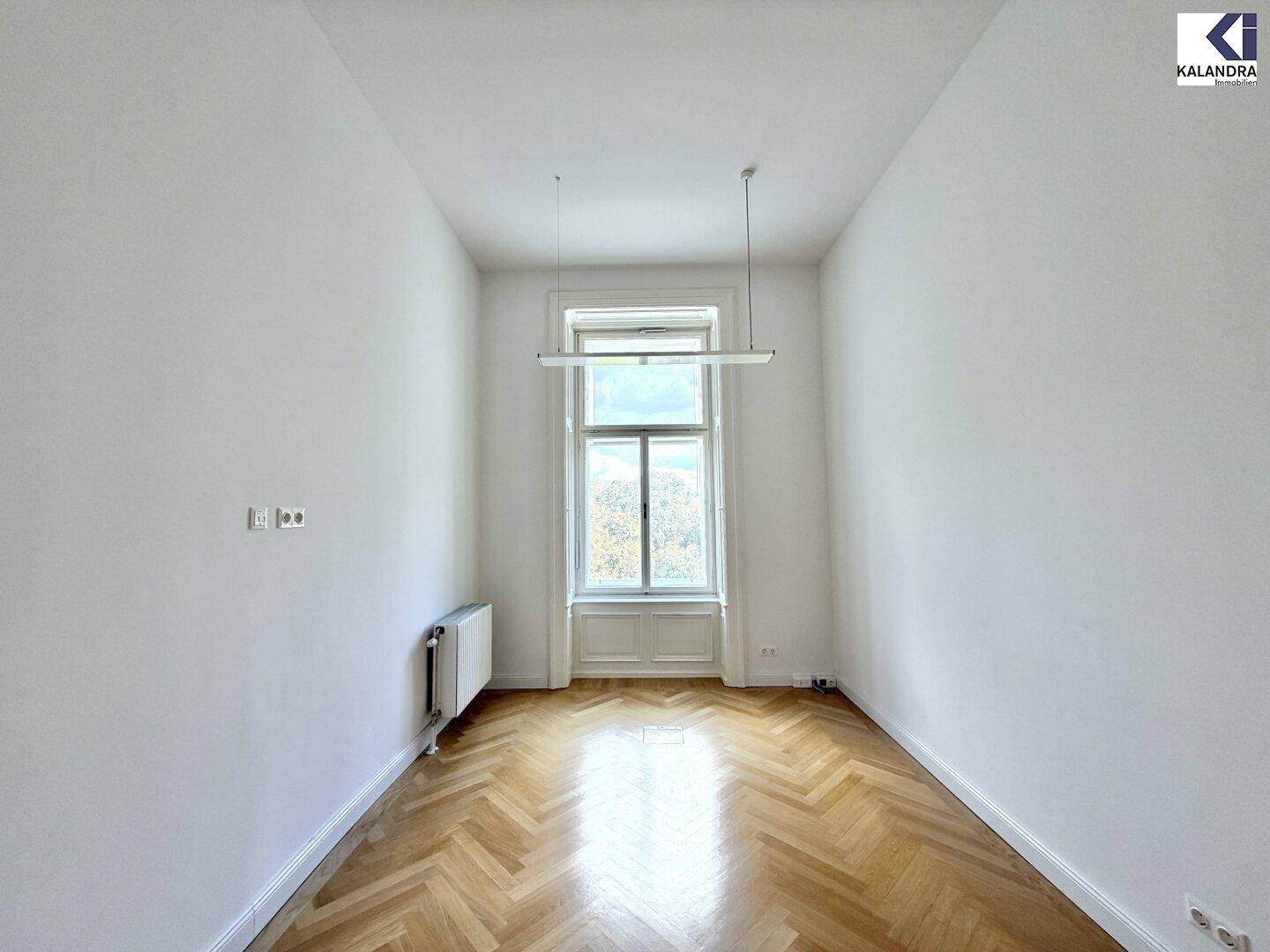 Heller, leerer Raum mit Fischgrätparkett und einem hohen Fenster, das viel Tageslicht hereinlässt.