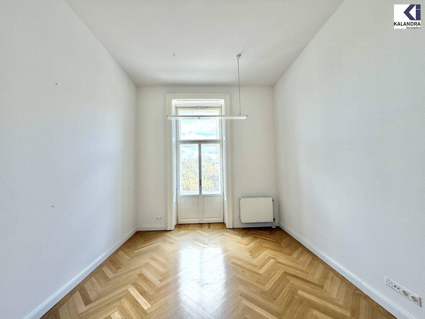 Helles Zimmer mit Fischgrätparkett, weißen Wänden und großem Fenster, das viel Tageslicht hereinlässt.