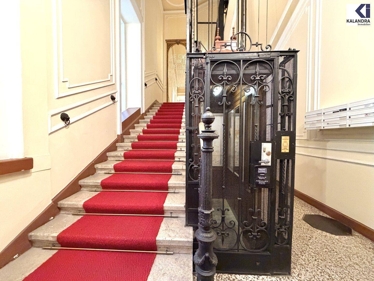 Historisches Treppenhaus mit rotem Teppich und einem kunstvoll verzierten Gitteraufzug.
