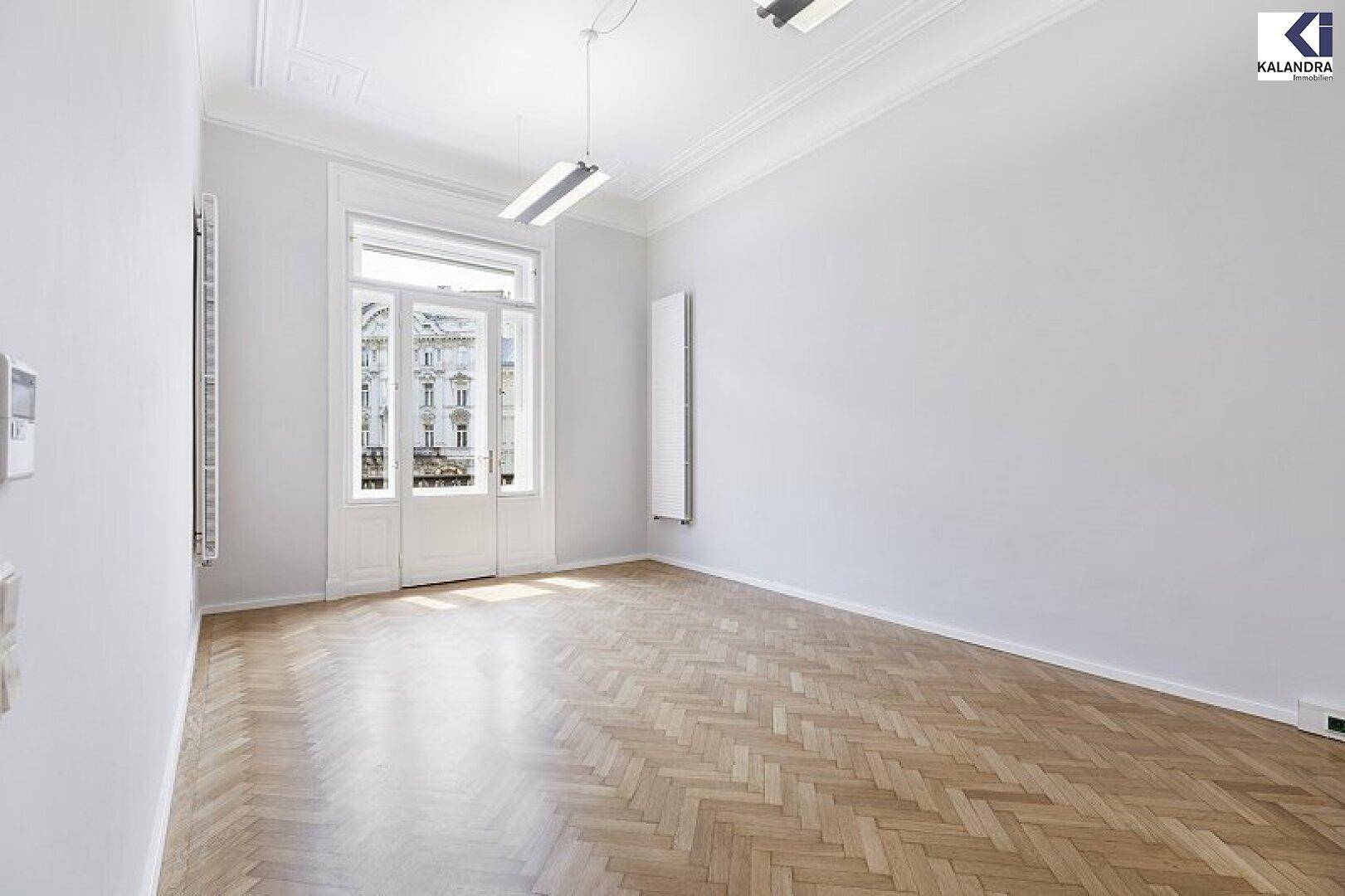 Geräumiges, leeres Zimmer mit Fischgrätparkett, hohen Decken und einem großen Fenster mit Blick nach draußen.
