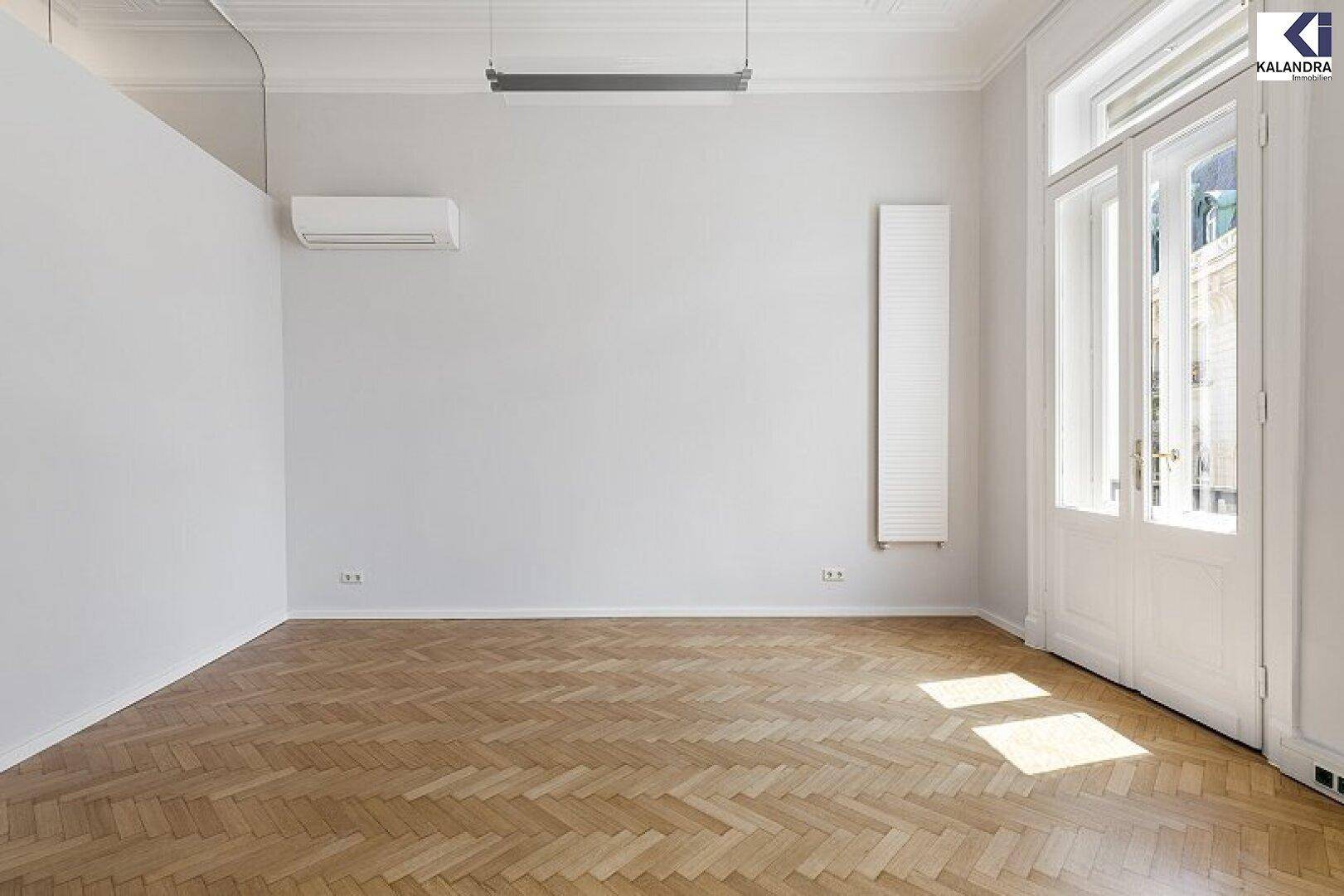 Helles Zimmer mit Fischgrätparkett, hohen Decken, Klimaanlage und einem großen Fenster, das viel Licht spendet.