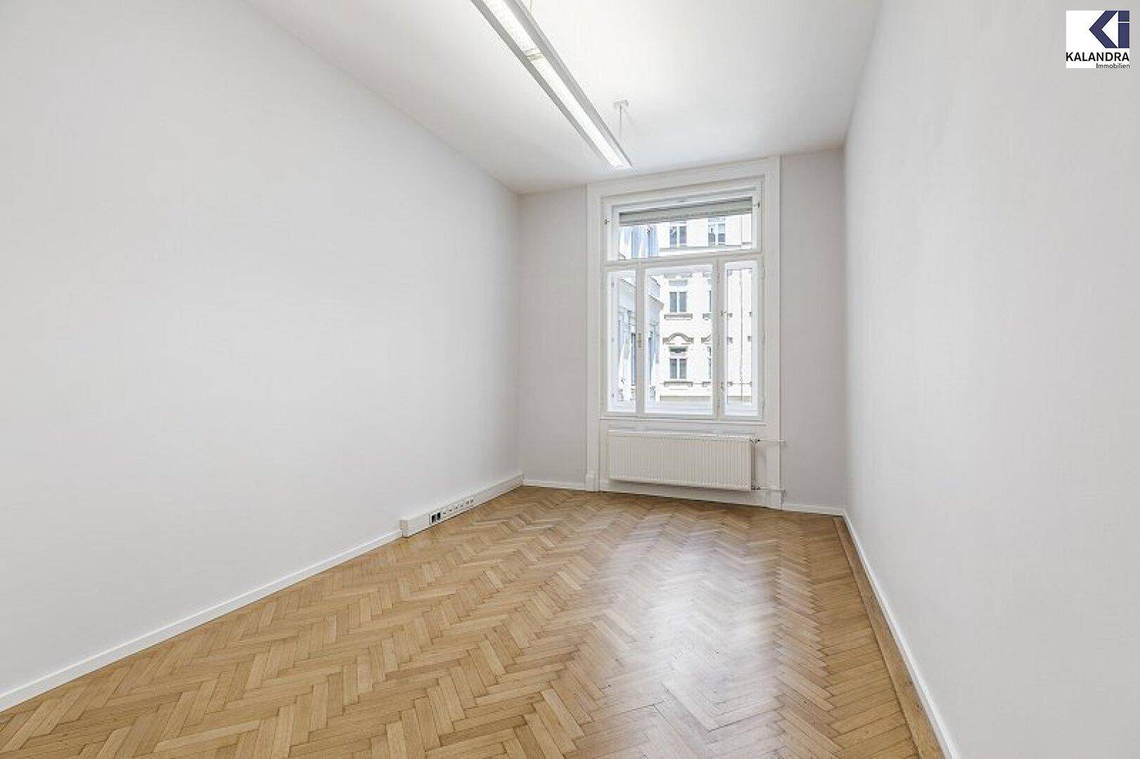Kleineres, helles Zimmer mit Fischgrätparkett und einem Fenster, das einen Blick auf die umliegenden Gebäude bietet.