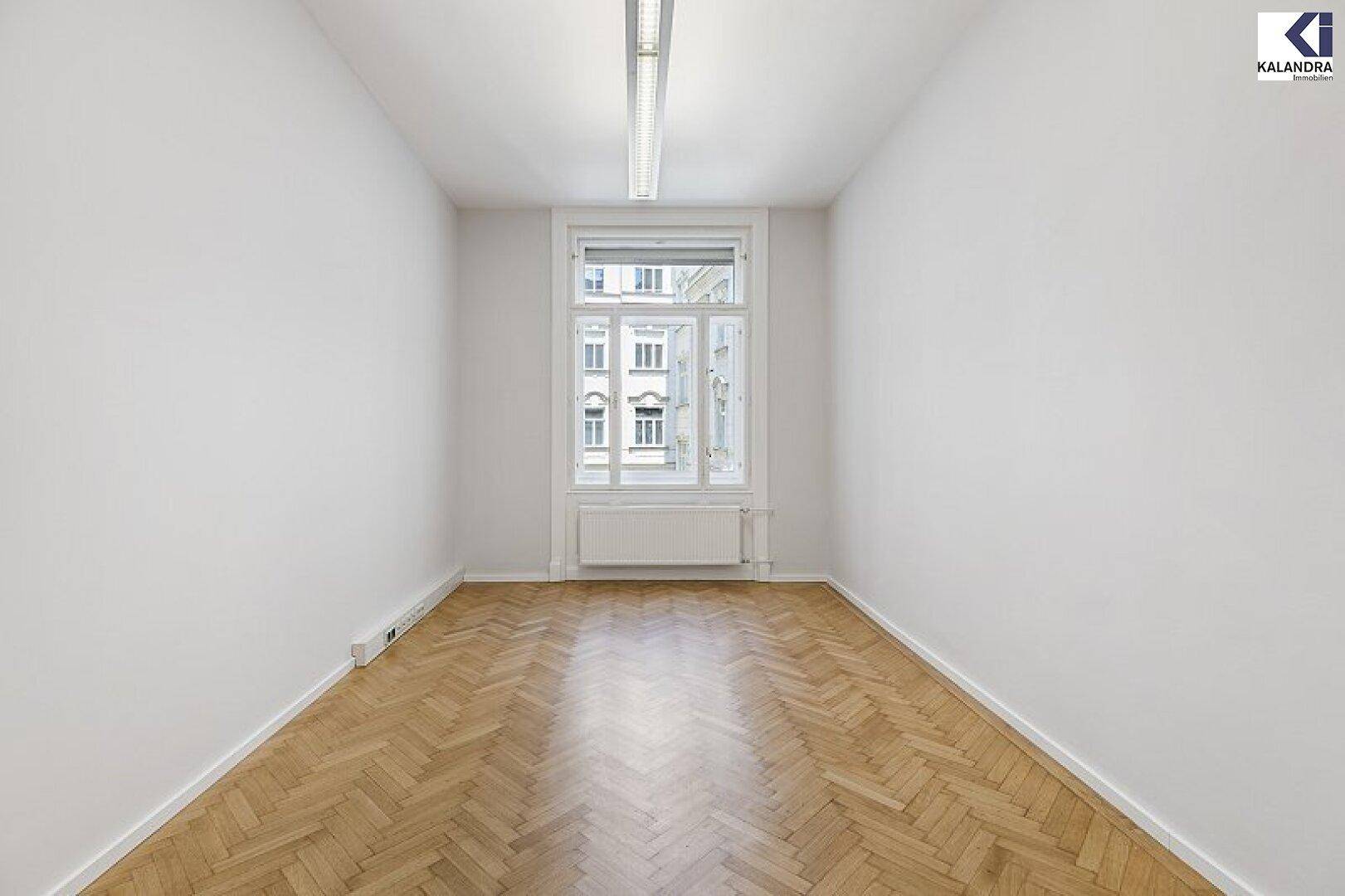 Kleineres, helles Zimmer mit Fischgrätparkett und einem Fenster, das einen Blick auf die umliegenden Gebäude bietet.