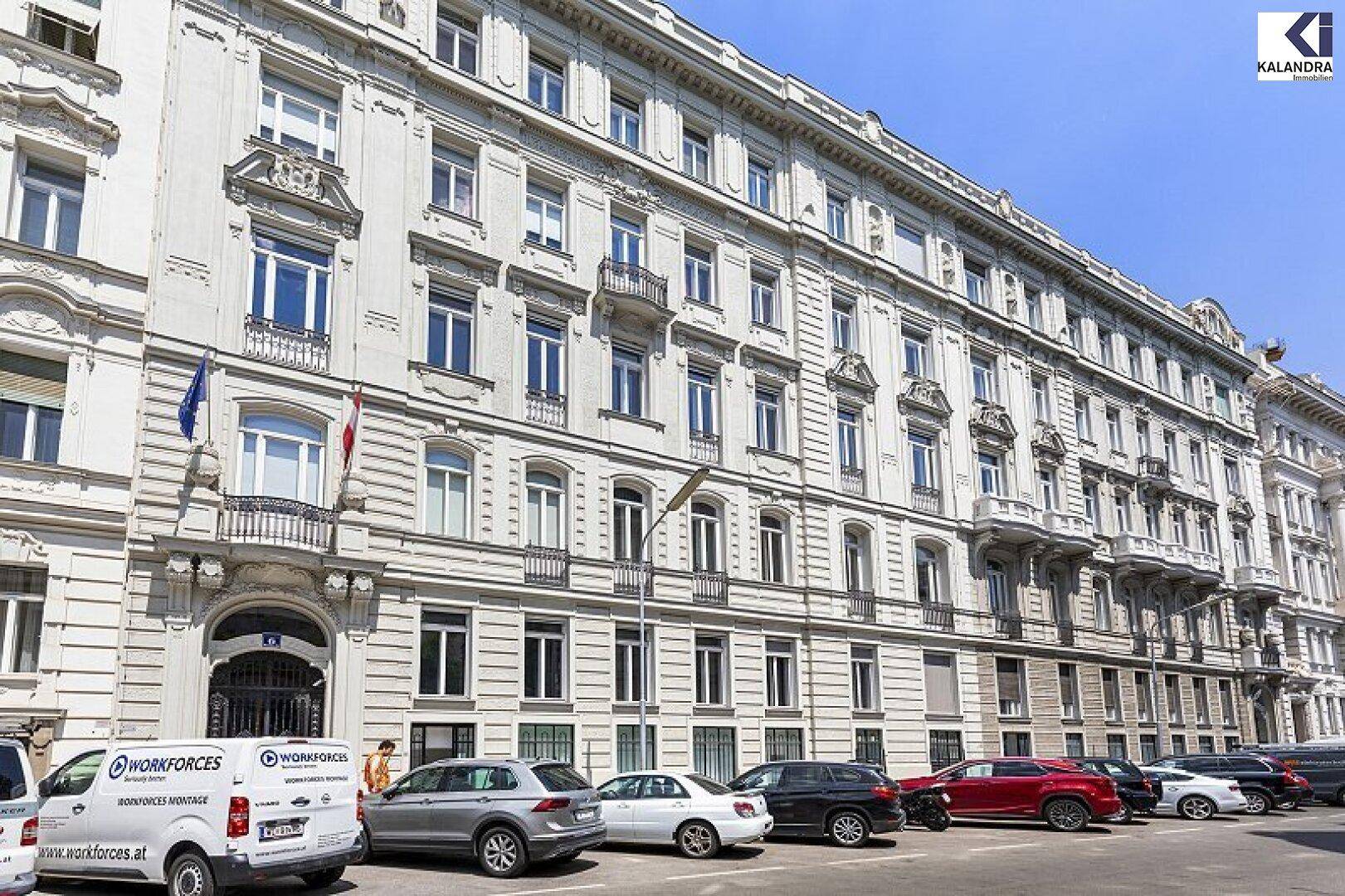 Lange Fassade eines mehrstöckigen Gebäudes mit zahlreichen Fenstern und geparkten Fahrzeugen davor.