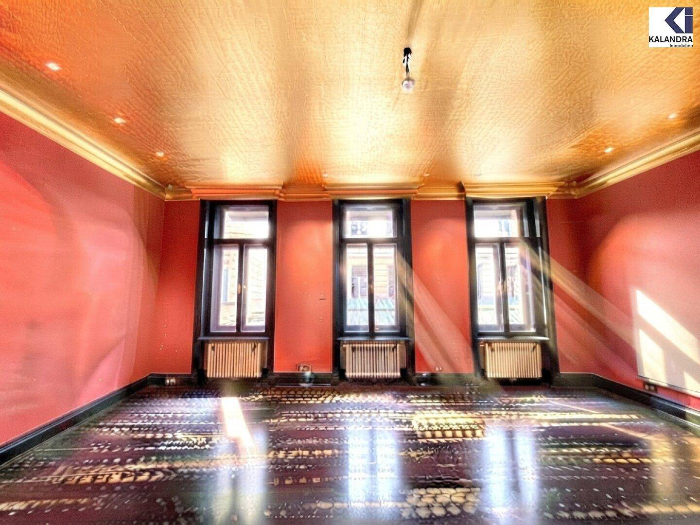 Helles Zimmer mit roten Wänden, goldener Decke und dunklem Fischgrätparkett, durchflutet von Sonnenlicht.