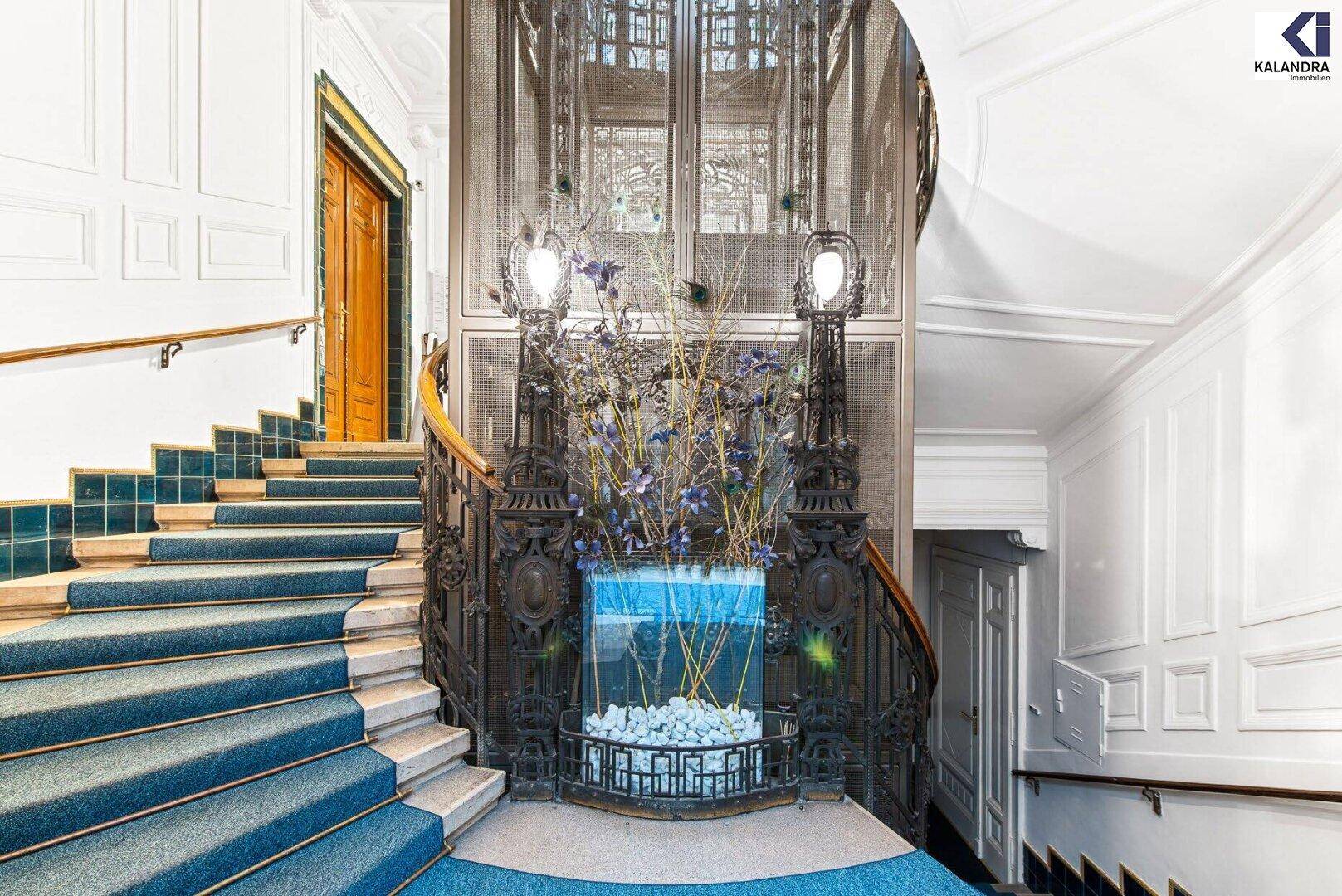 Eindrucksvolles Treppenhaus mit kunstvollem Aufzug und blauen Teppichstufen.