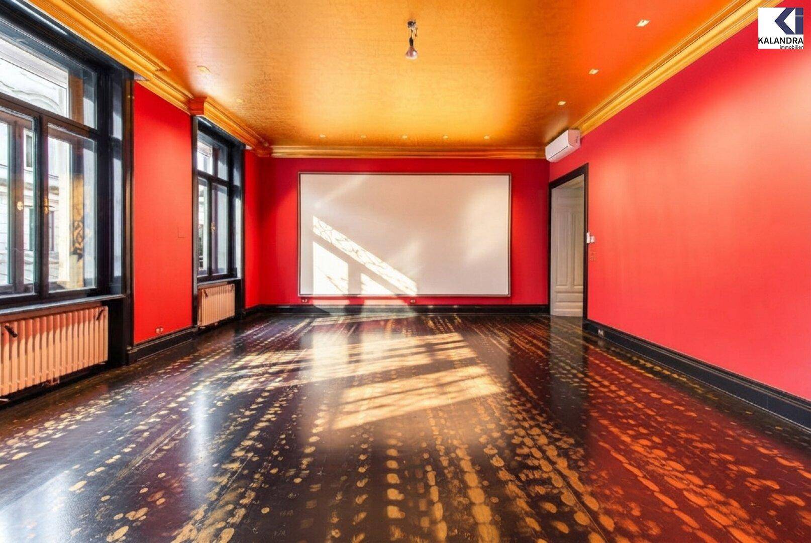 Großes Zimmer mit roter Wandfarbe, goldener Decke und dunklem Fischgrätparkett, inklusive Projektionsfläche.
