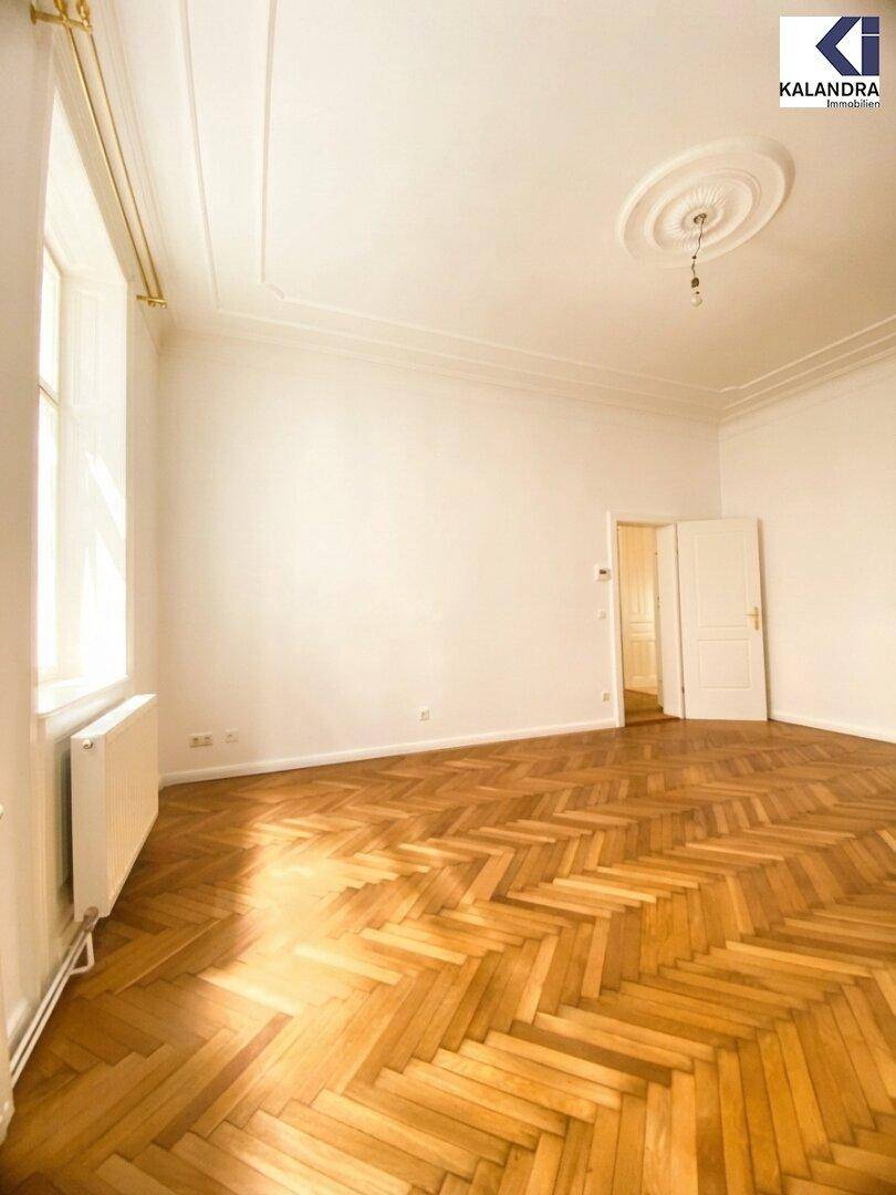 Heller Wohnraum mit klassischem Fischgrätparkett, hohen Decken und einer Tür, die zu einem weiteren Zimmer führt.