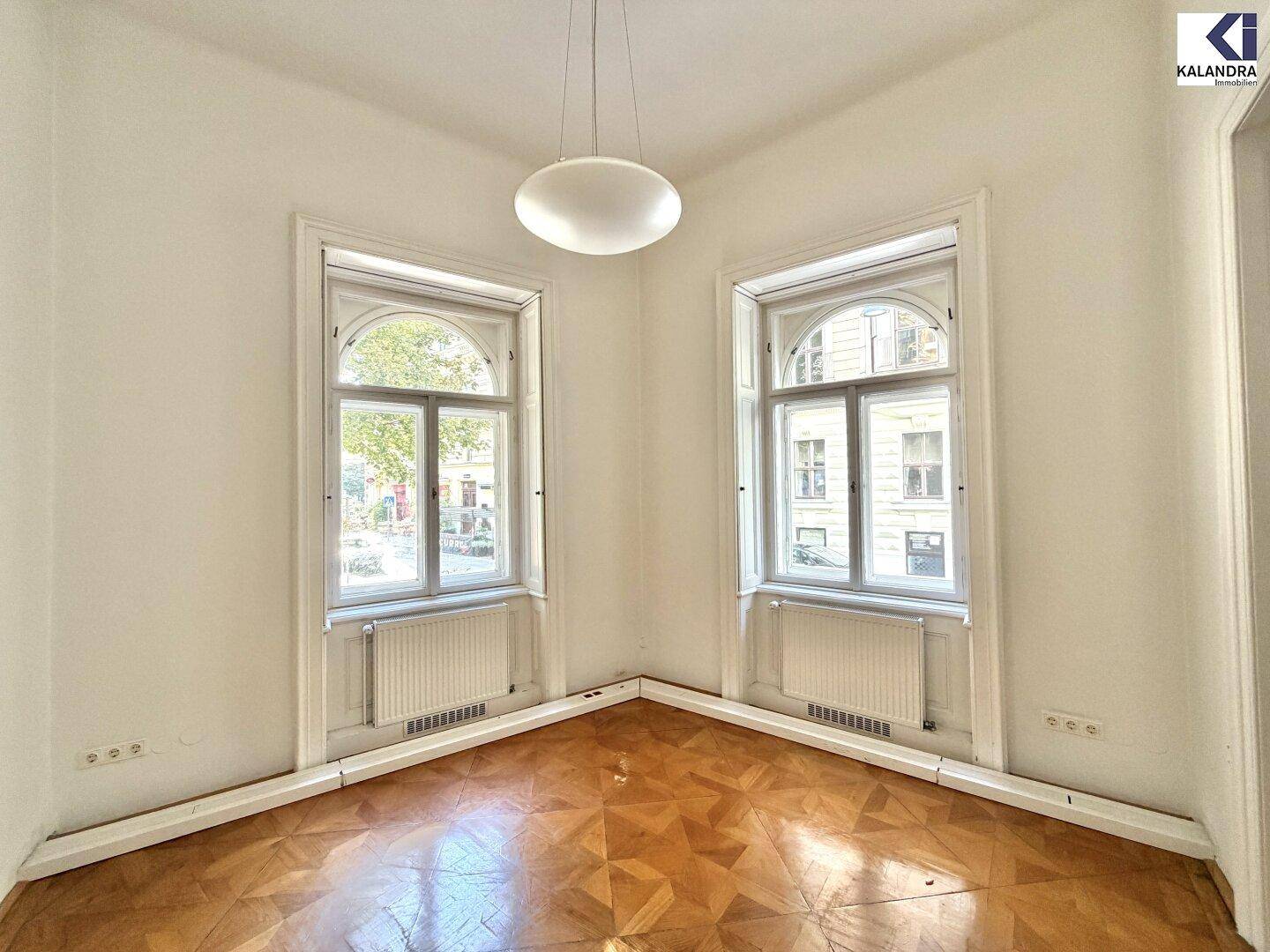 Gemütliche Ecke eines Zimmers mit zwei hohen Fenstern, die viel natürliches Licht hereinlassen, und Parkettboden.