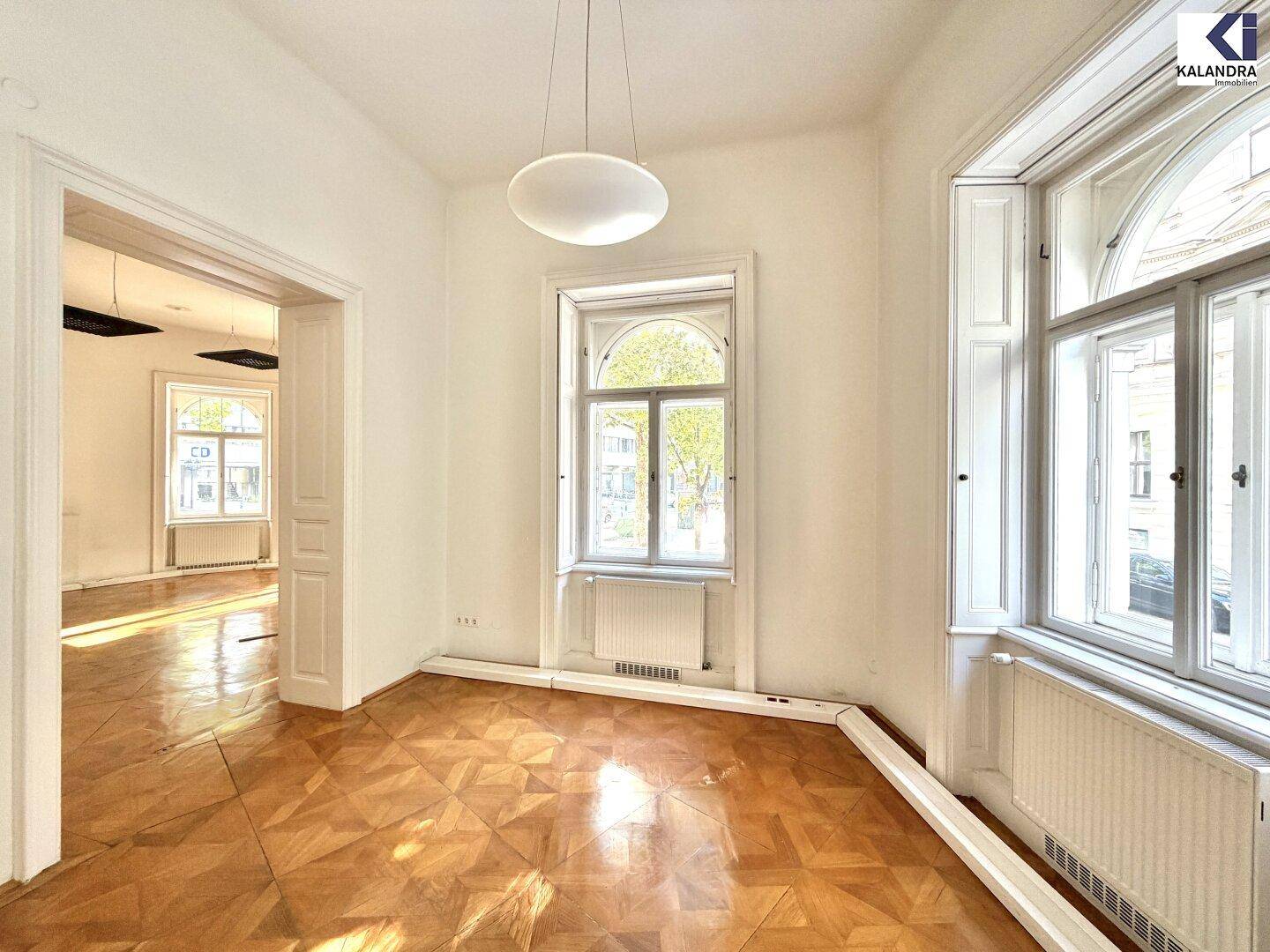 Helles Zimmer mit großem Fenster und Blick in einen angrenzenden Raum, ausgestattet mit Parkettboden.