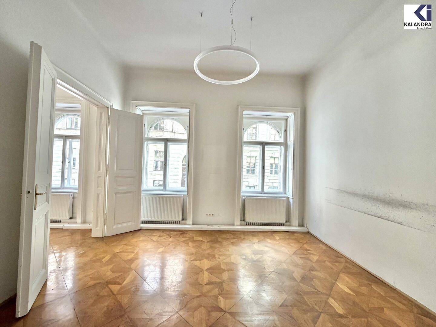 Geräumiges Zimmer mit Parkettboden, hohen Decken und großen Fenstern, die für eine helle Atmosphäre sorgen.