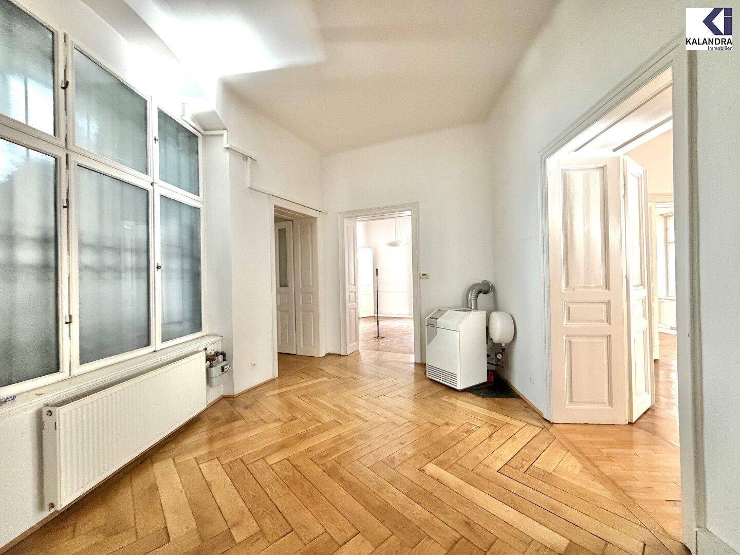 Heller Flur mit Fischgrätparkett, großen Fenstern und einem Heizgerät, der zu weiteren Räumen führt.