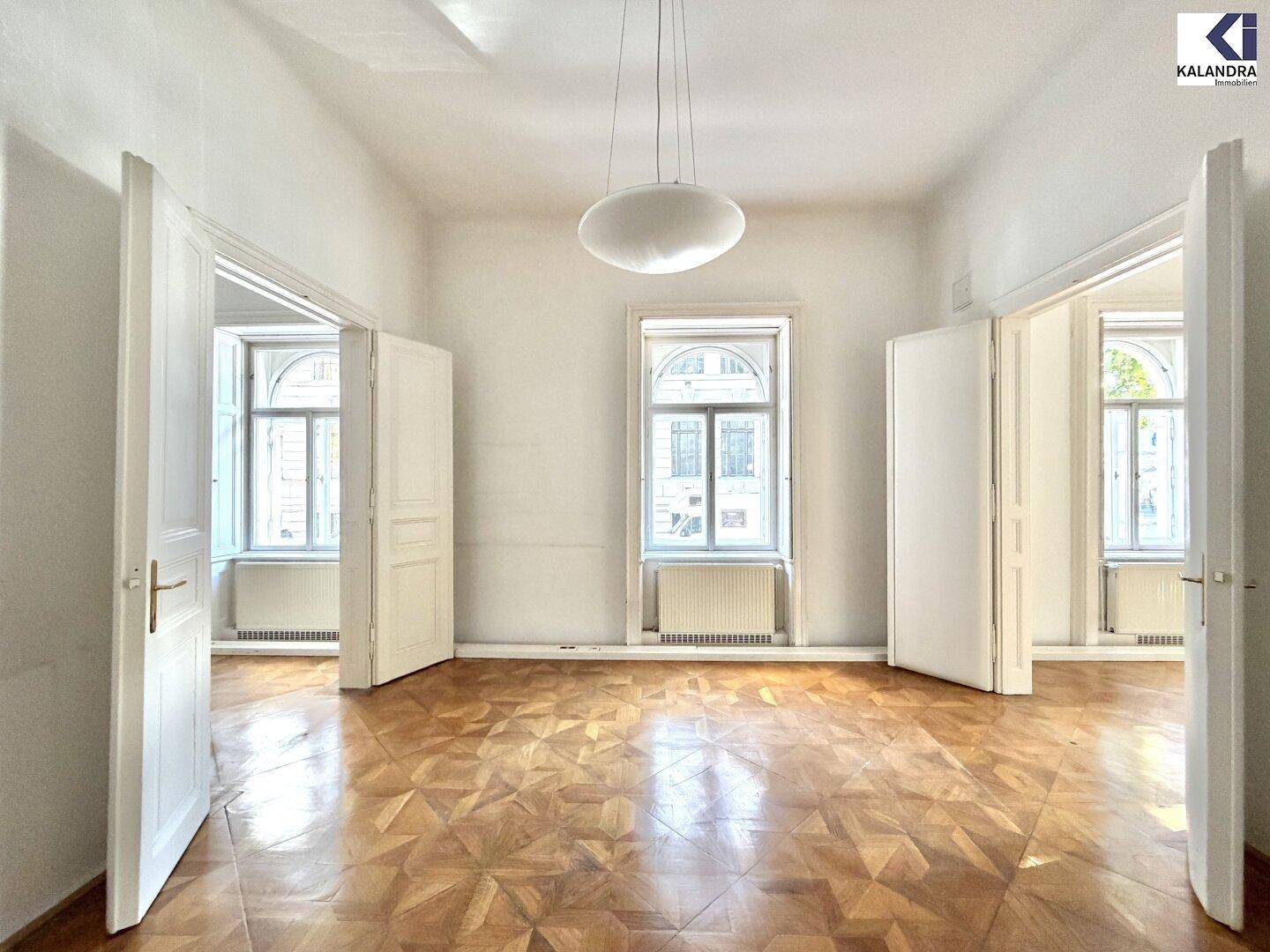 Klassisches Zimmer mit Parkettboden, hohen Fenstern und einer modernen Deckenleuchte, die den Raum erhellt.