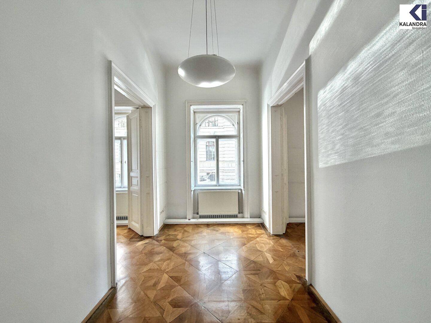 Langer Flur mit Fischgrätparkett und Zugang zu mehreren Zimmern, hell beleuchtet durch ein Fenster.