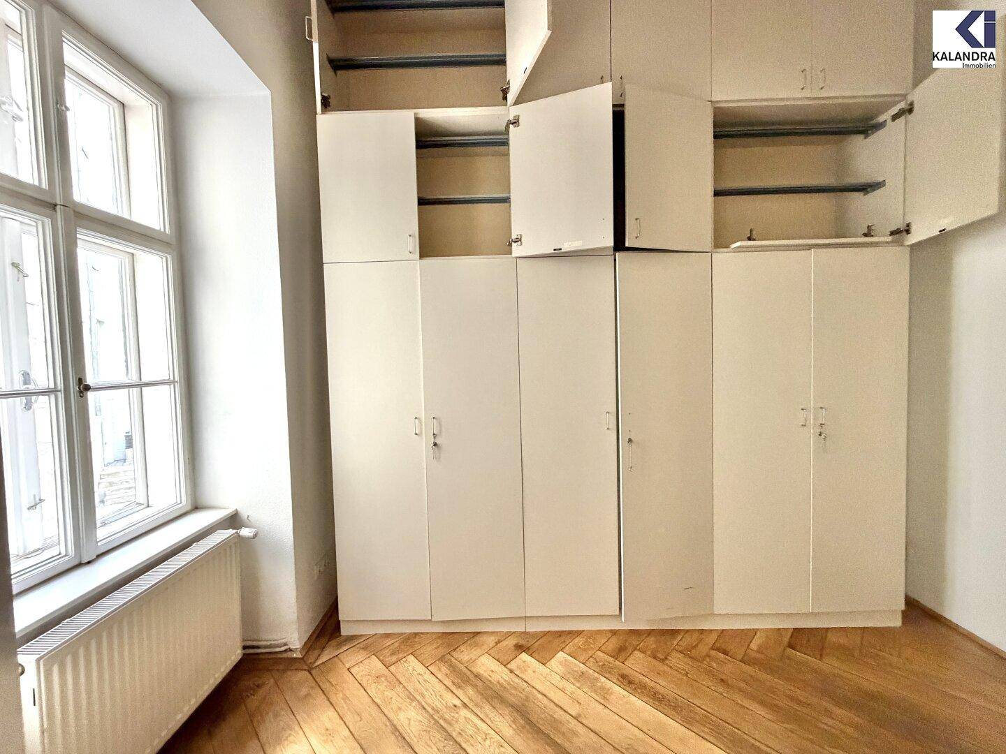 Helles Zimmer mit hohen Einbauschränken und Parkettboden neben einem großen Fenster, ideal als Schlafzimmer.
