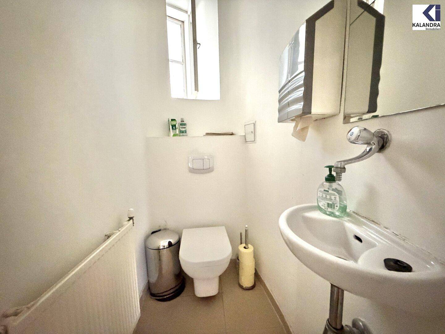 Separate Toilette mit Waschbecken, Fenster und Wand-WC, in einem hellen Raum mit Fliesenboden.
