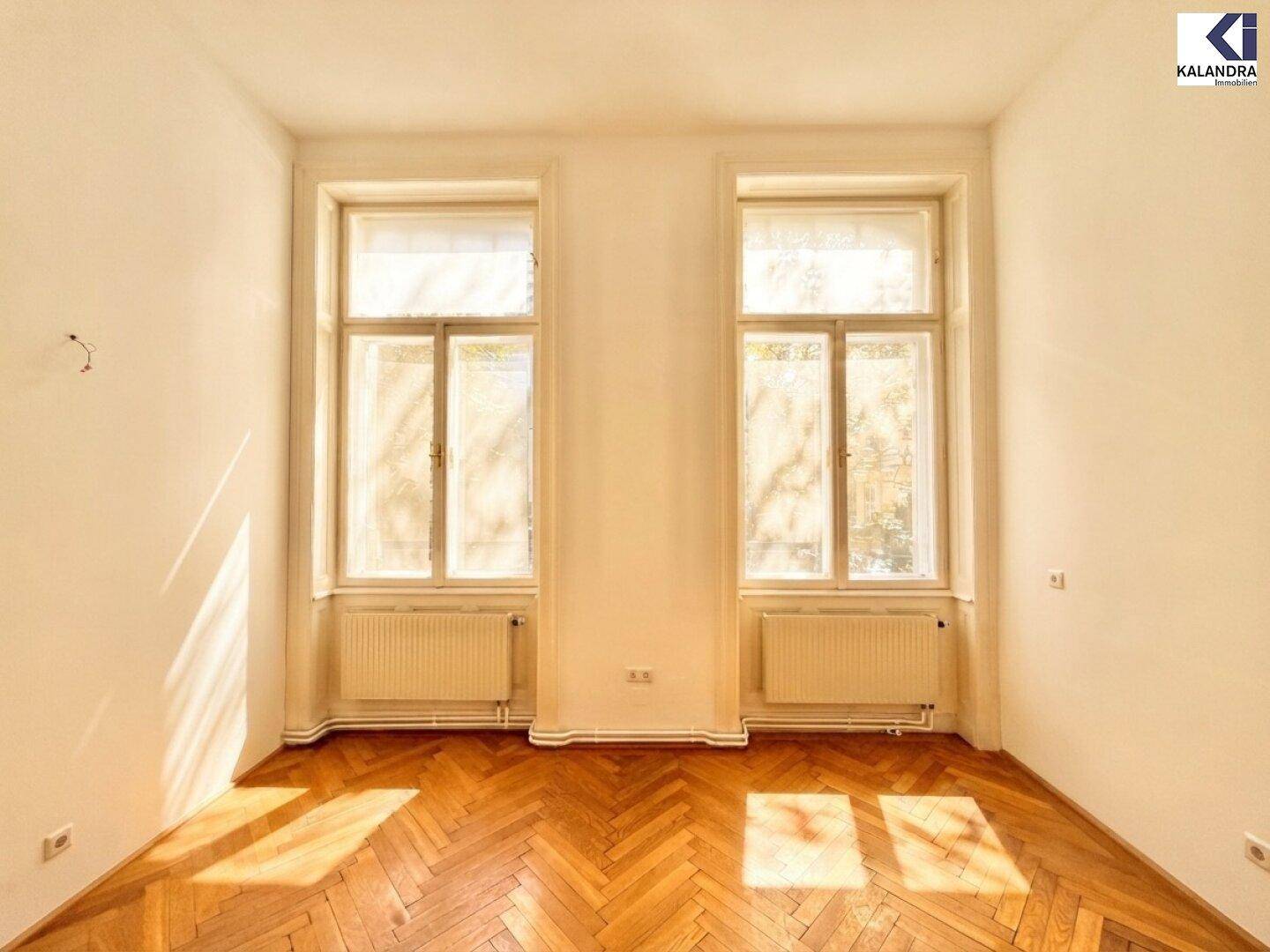 Sonniger Raum mit Fischgrätparkett und zwei hohen Fenstern, die viel Licht hereinlassen.