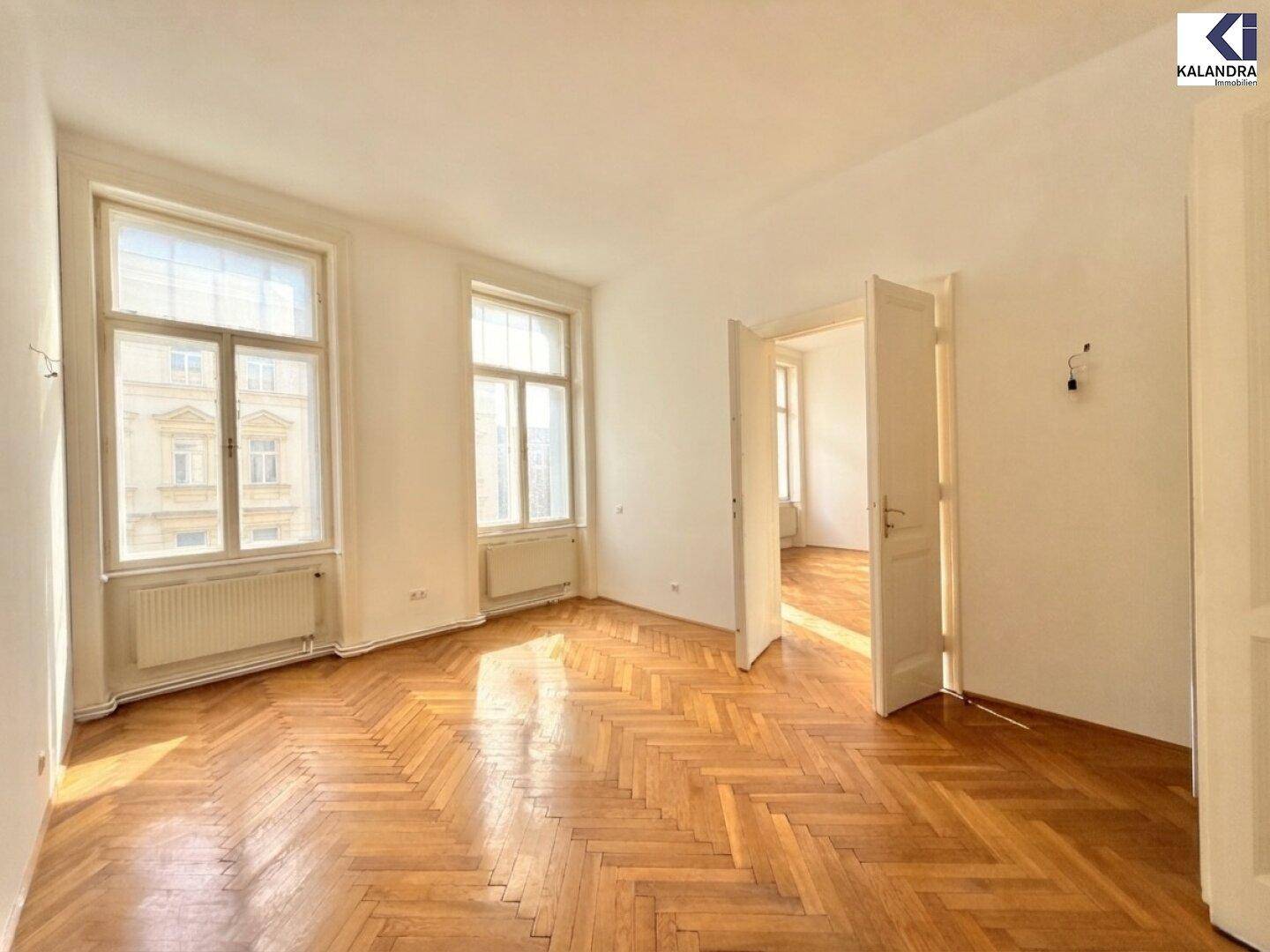 Großes Wohnzimmer mit Fischgrätparkett, hohen Decken und Zugang zu weiteren Räumen.