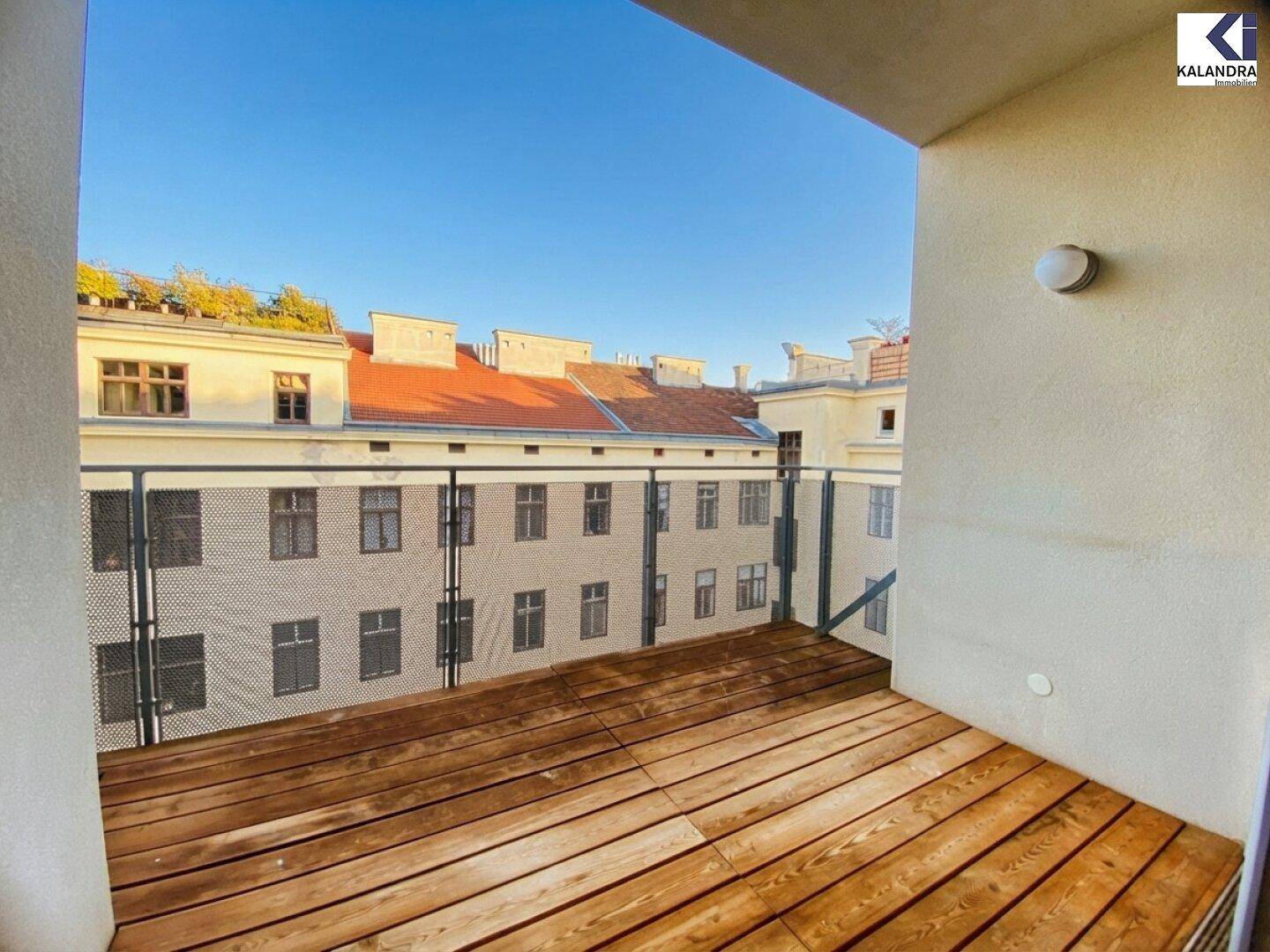 Sonniger Balkon mit Holzboden und Blick auf die umliegenden Gebäude im Innenhof.