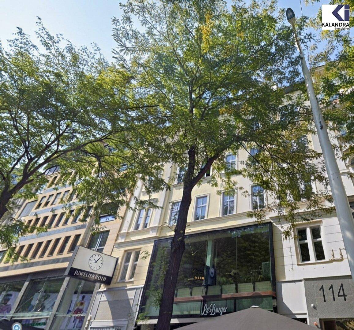Fassade eines Gebäudes an einer belebten Straße mit Geschäften und Bäumen.