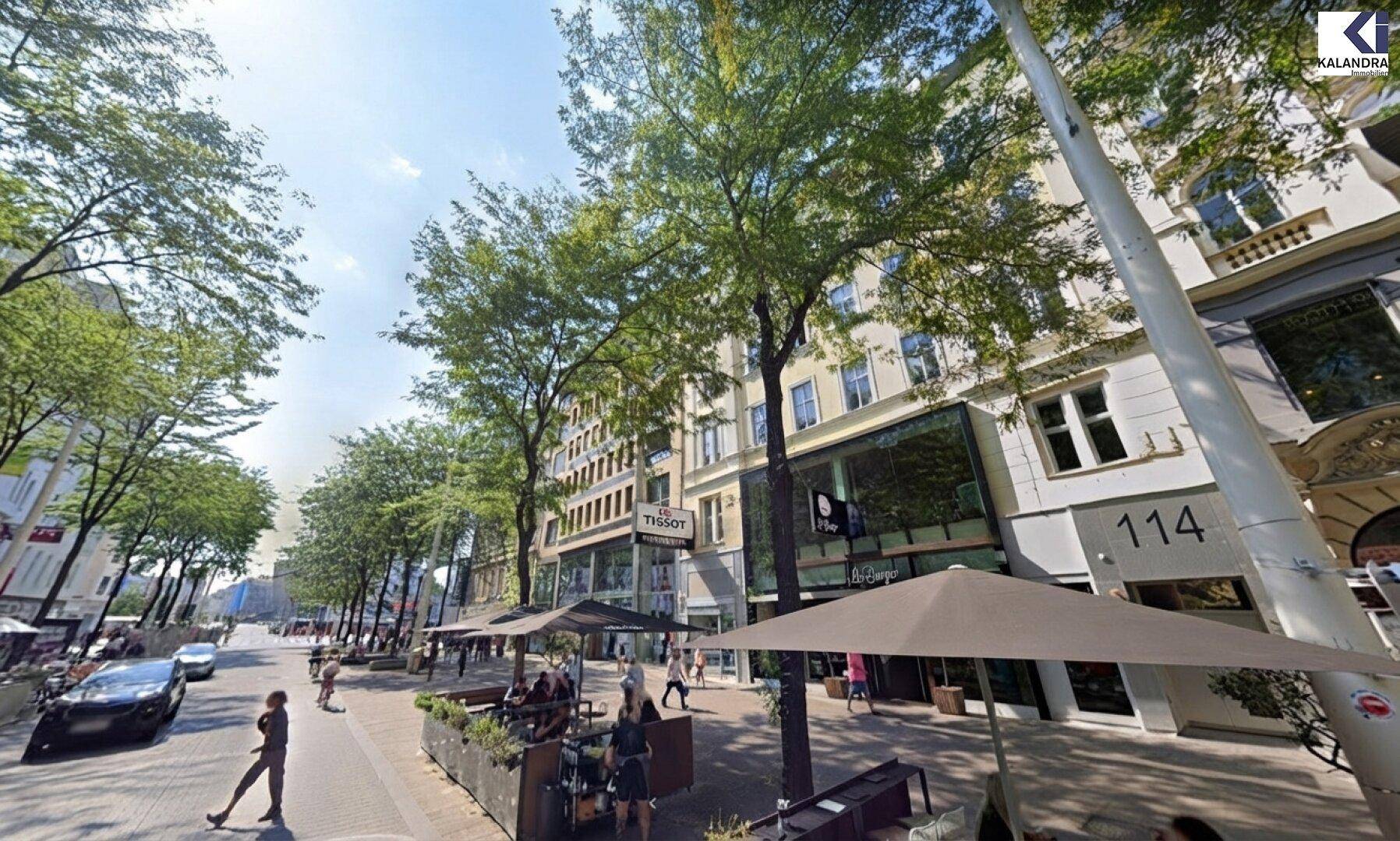 Belebtes Stadtbild mit Geschäften, Restaurants und Bäumen entlang einer breiten Straße.