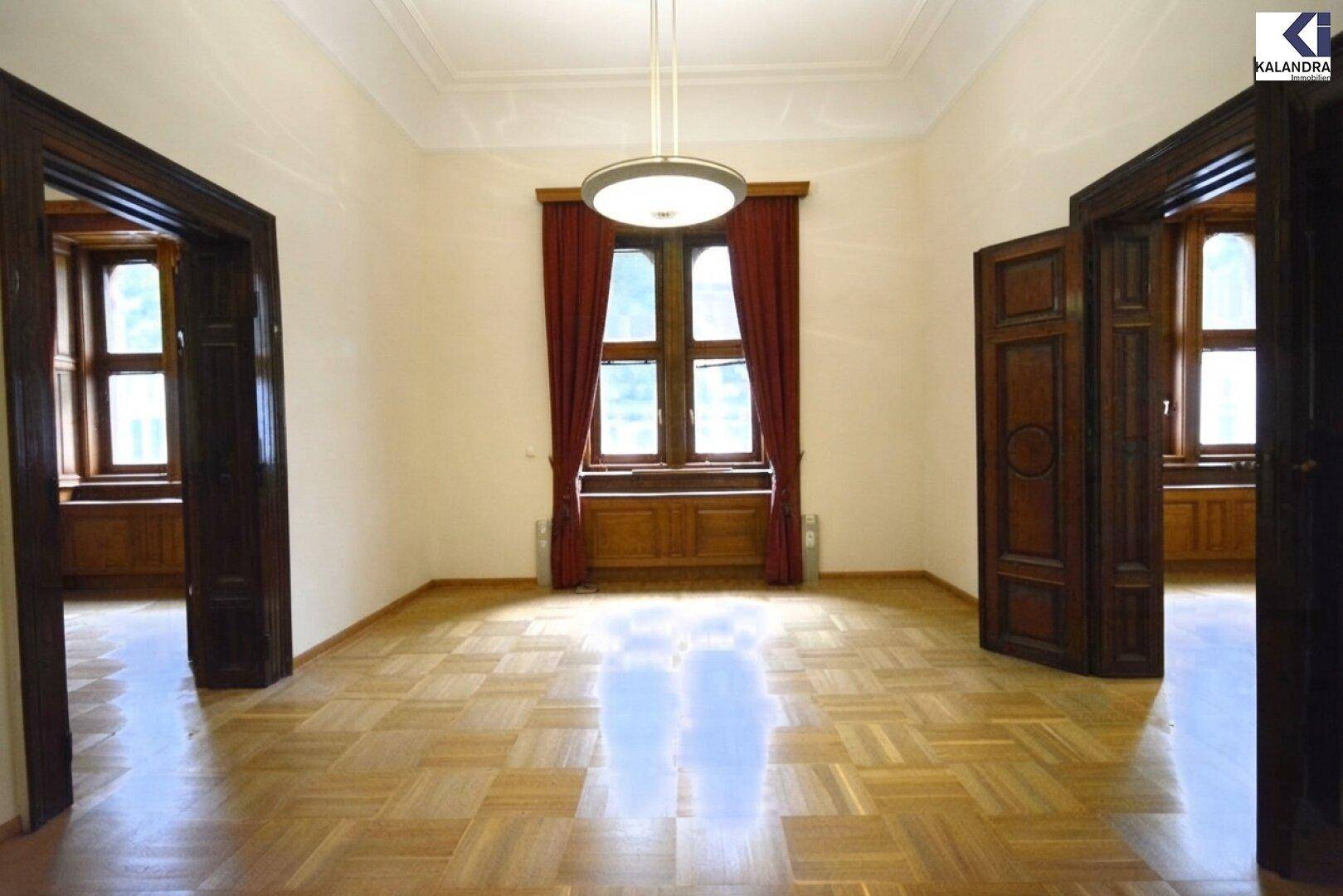 Geräumiger Wohnraum mit klassischem Parkettboden, hohen Decken und zwei Holztüren zu angrenzenden Räumen.