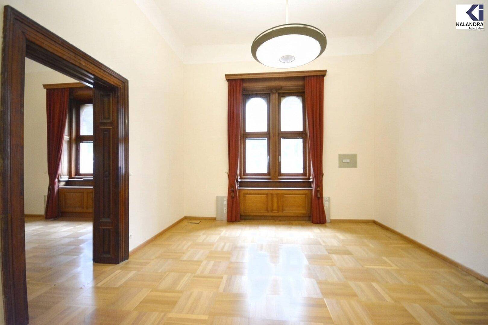 Klassischer Raum mit hellem Parkettboden, hohen Decken und einem großen Fenster mit roten Vorhängen.