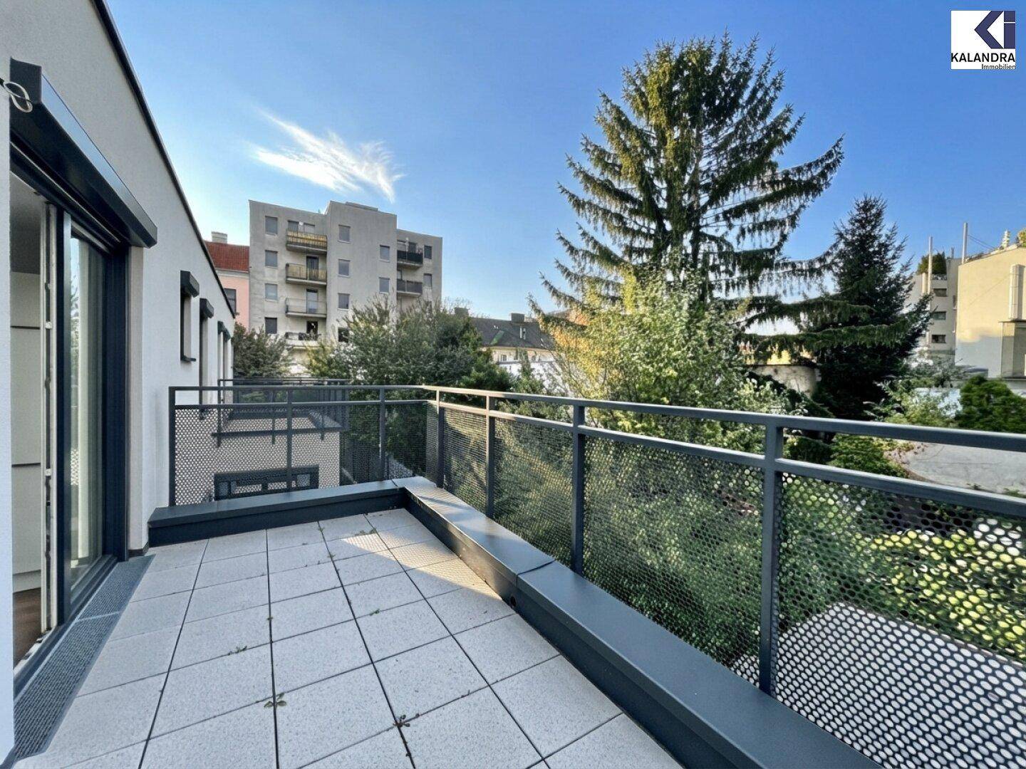 Geräumige Außenterrasse mit Fliesenboden und Blick auf die umliegenden Gebäude und Bäume.