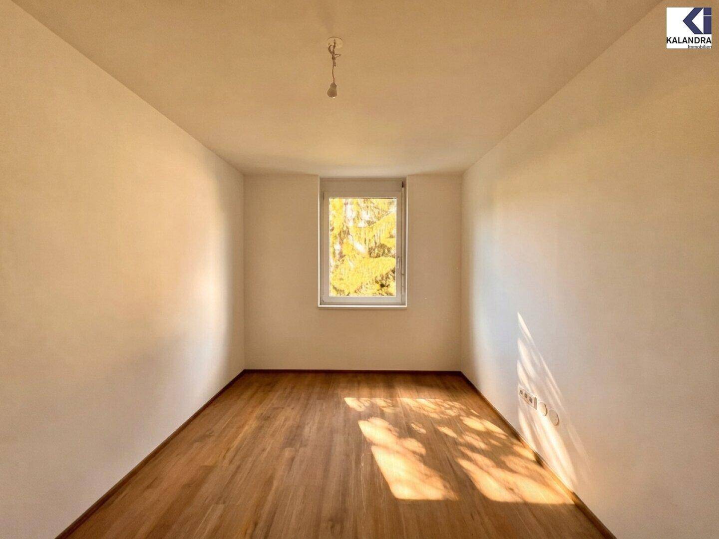 Leeres Zimmer mit hellem Holzboden und einem Fenster, das viel Tageslicht hereinlässt.