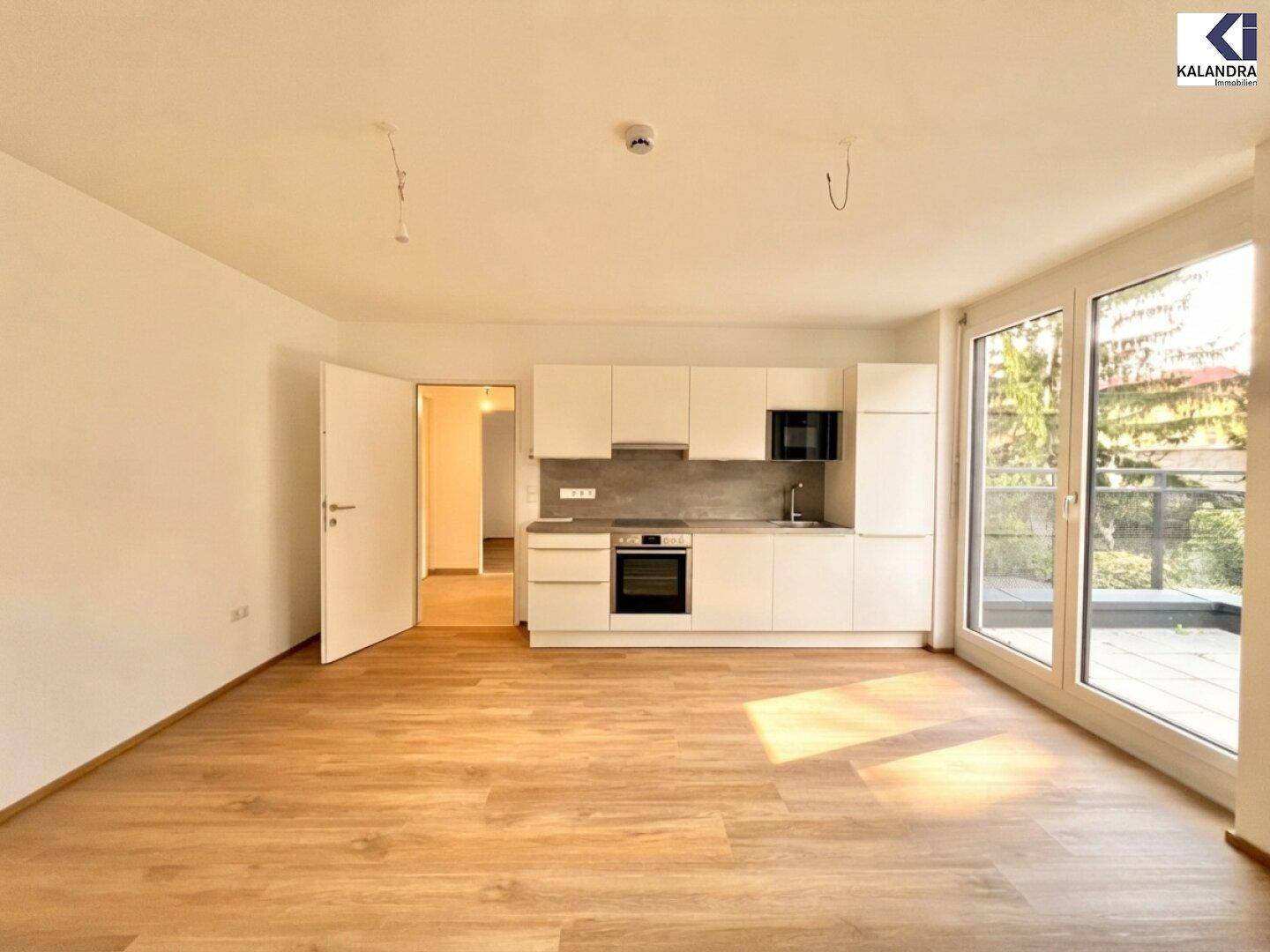 Offener Wohnbereich mit moderner Einbauküche und hellem Holzboden, der zum Balkon führt.