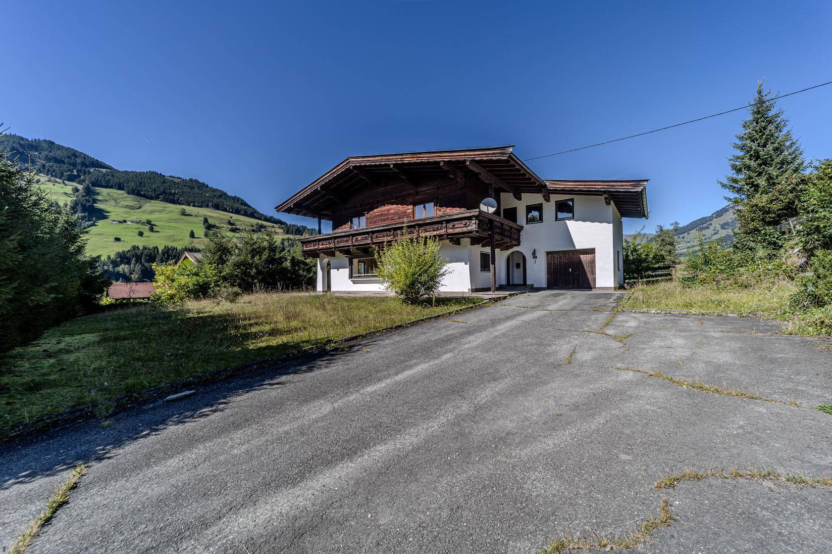 Traditionelles Haus mit Holzbalkon und langer Auffahrt, umgeben von grünen Hügeln unter blauem Himmel.