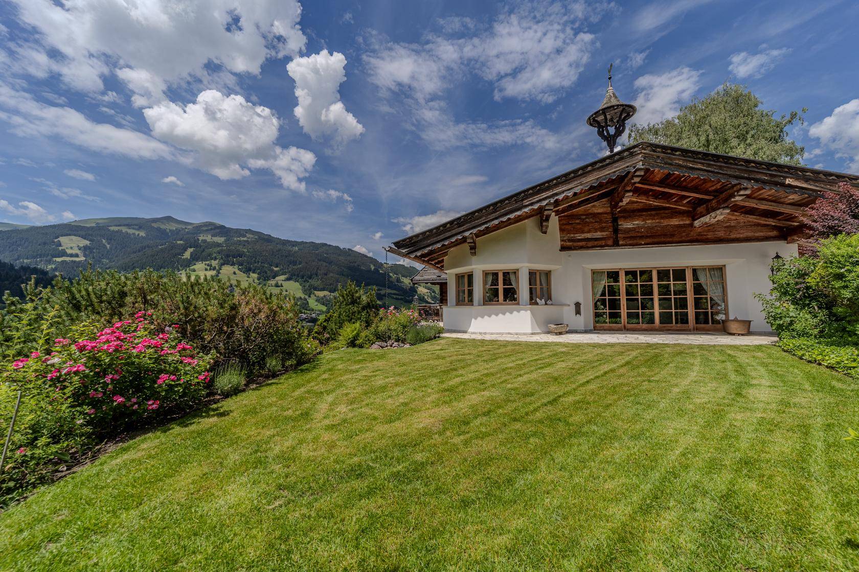 Idyllisches Landhaus mit weitläufigem Rasen und blühenden Sträuchern, umgeben von grünen Bergen.