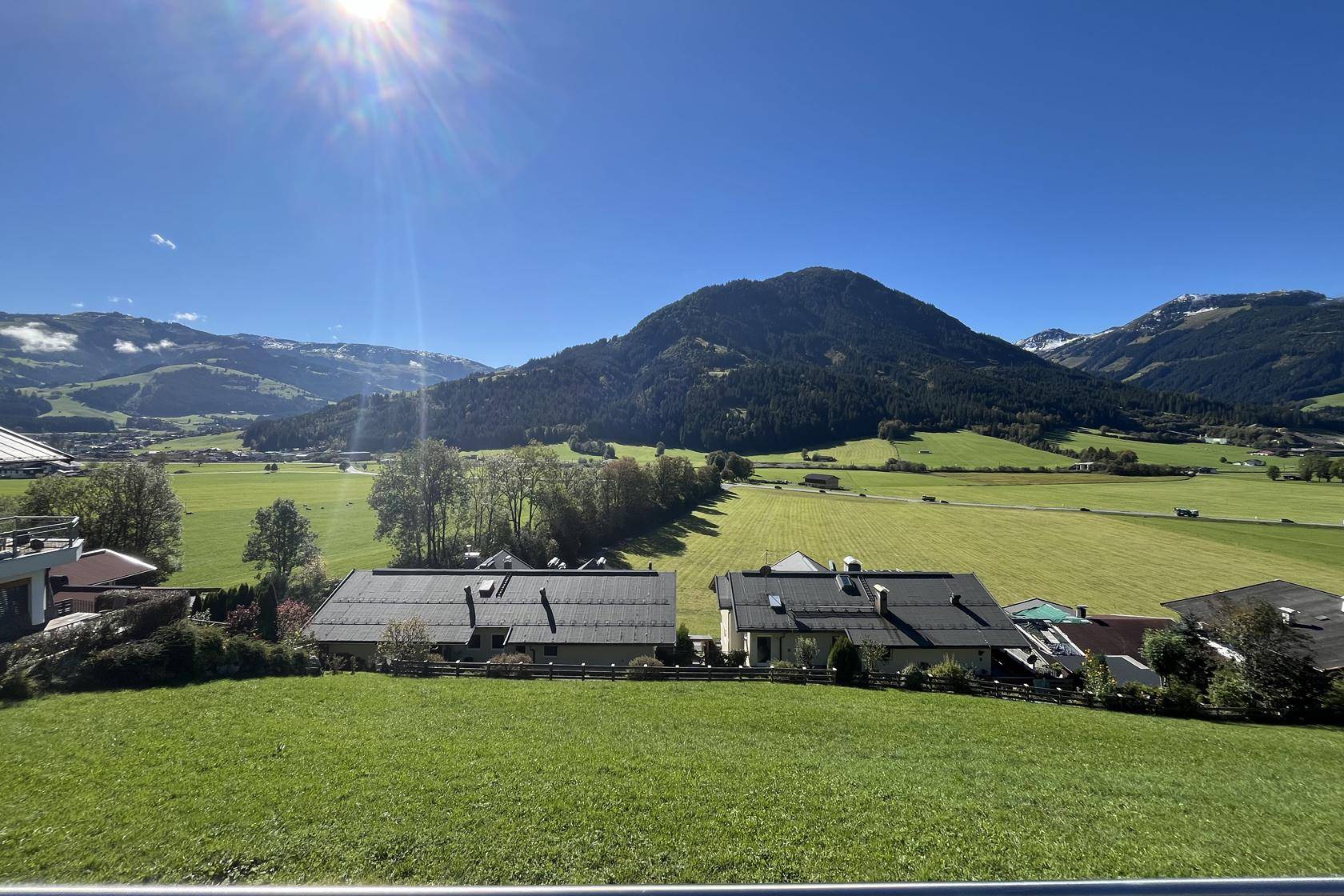 Sonniger Ausblick auf die weite Natur mit grünen Wiesen und bewaldeten Bergen unter strahlend blauem Himmel.