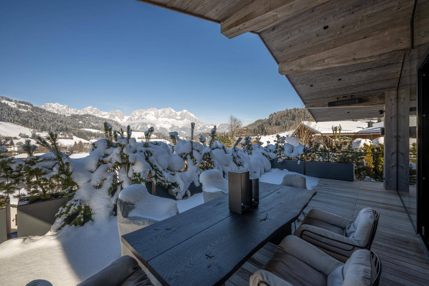 Sonnige Terrasse mit Holzboden, umgeben von verschneiten Bäumen und beeindruckendem Bergblick.