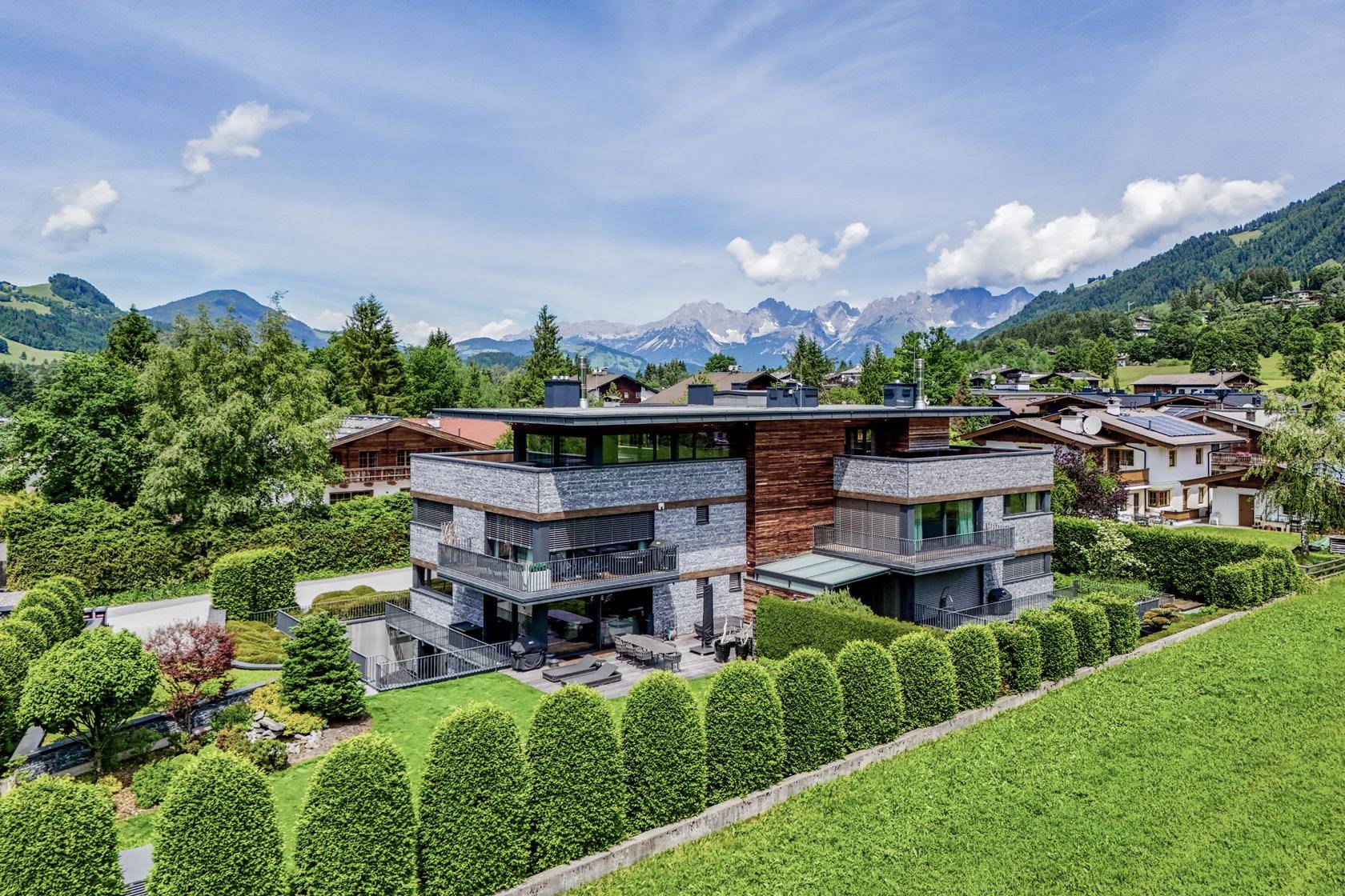 Moderne Villa mit Steinfassade und Holzdetails, umgeben von gepflegtem Garten und beeindruckendem Alpenpanorama.