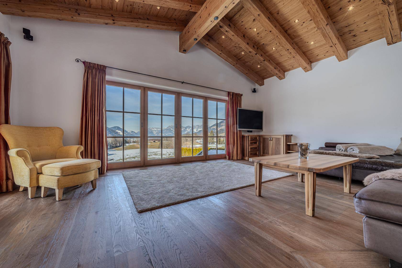 Helles Wohnzimmer mit Holzbalkendecke, Parkettboden und Panoramafenstern mit winterlichem Bergblick.