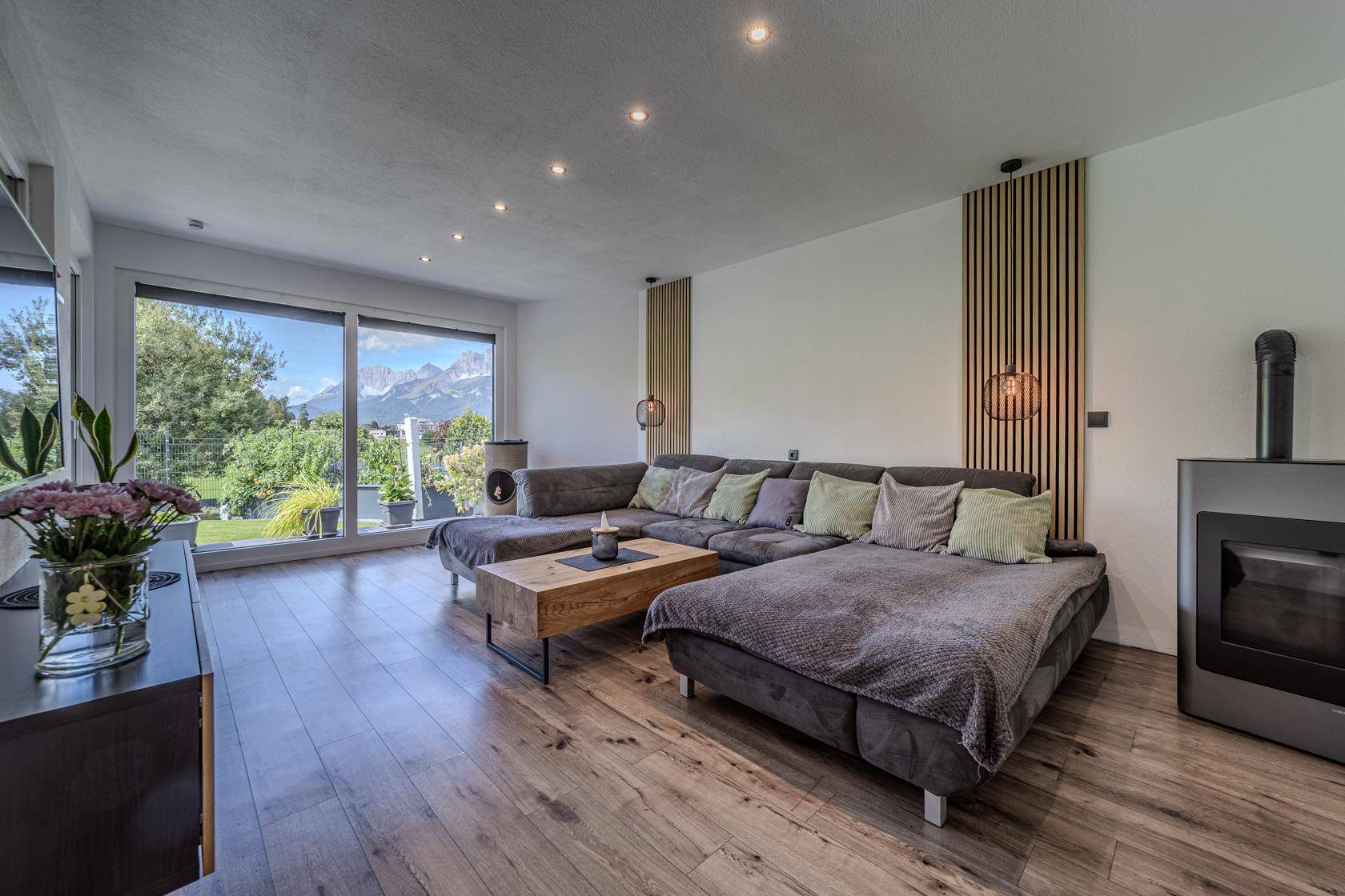 Helles Wohnzimmer mit großem Sofa, Holzboden und bodentiefen Fenstern mit Bergblick.
