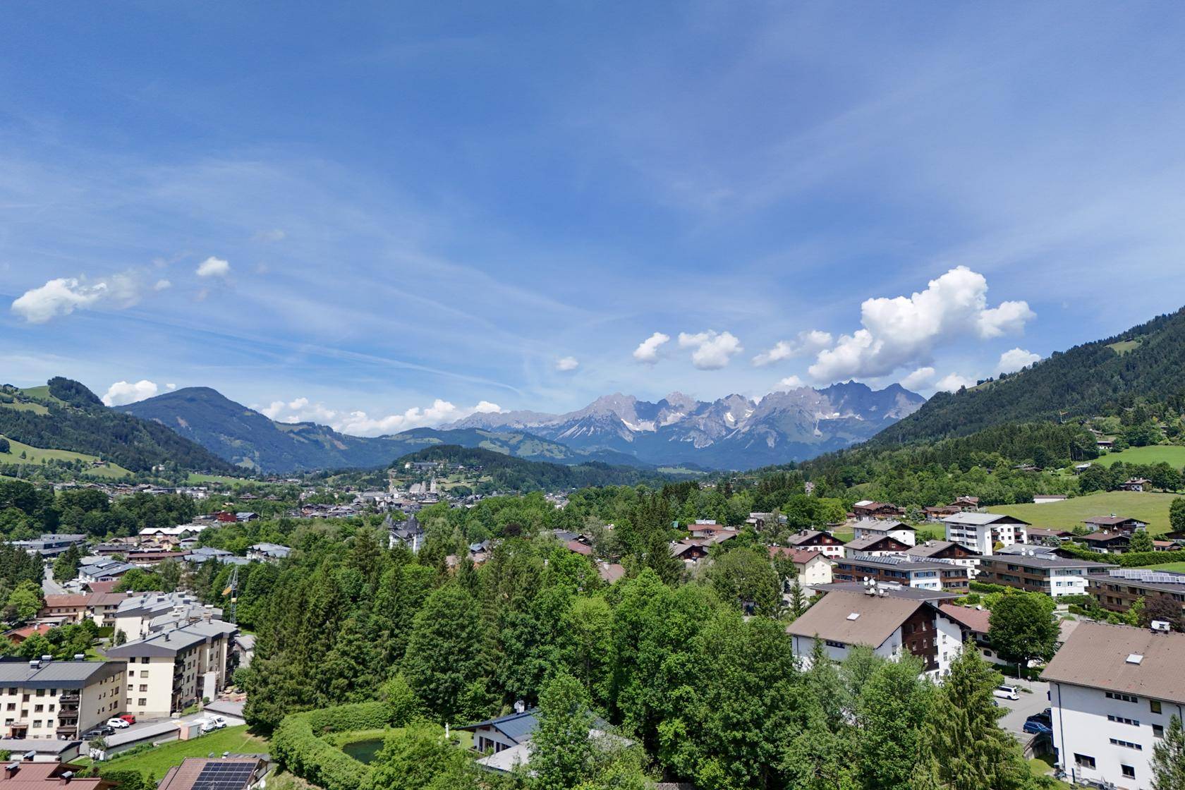 Panoramablick über eine malerische Stadt im Tal, umgeben von grünen Bergen und blauem Himmel.