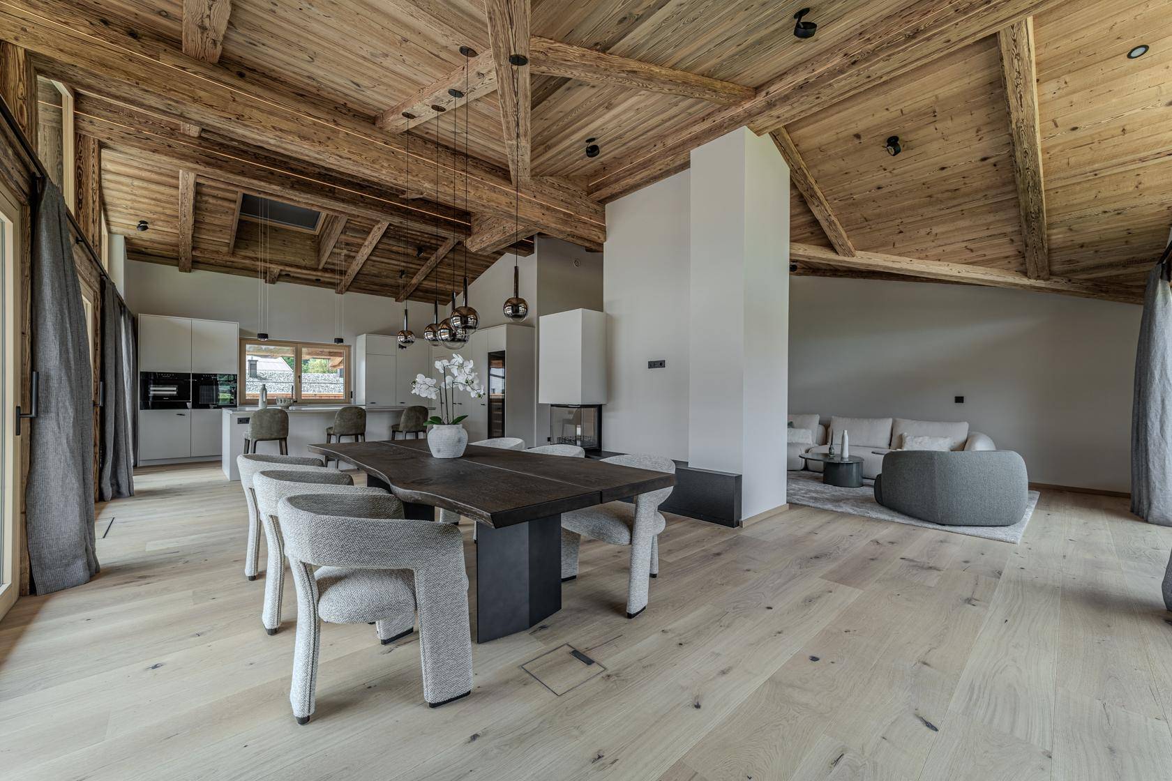 Geräumiger offener Wohn- und Essbereich mit Holzbalkendecke, Kamin und moderner Küche im Hintergrund.