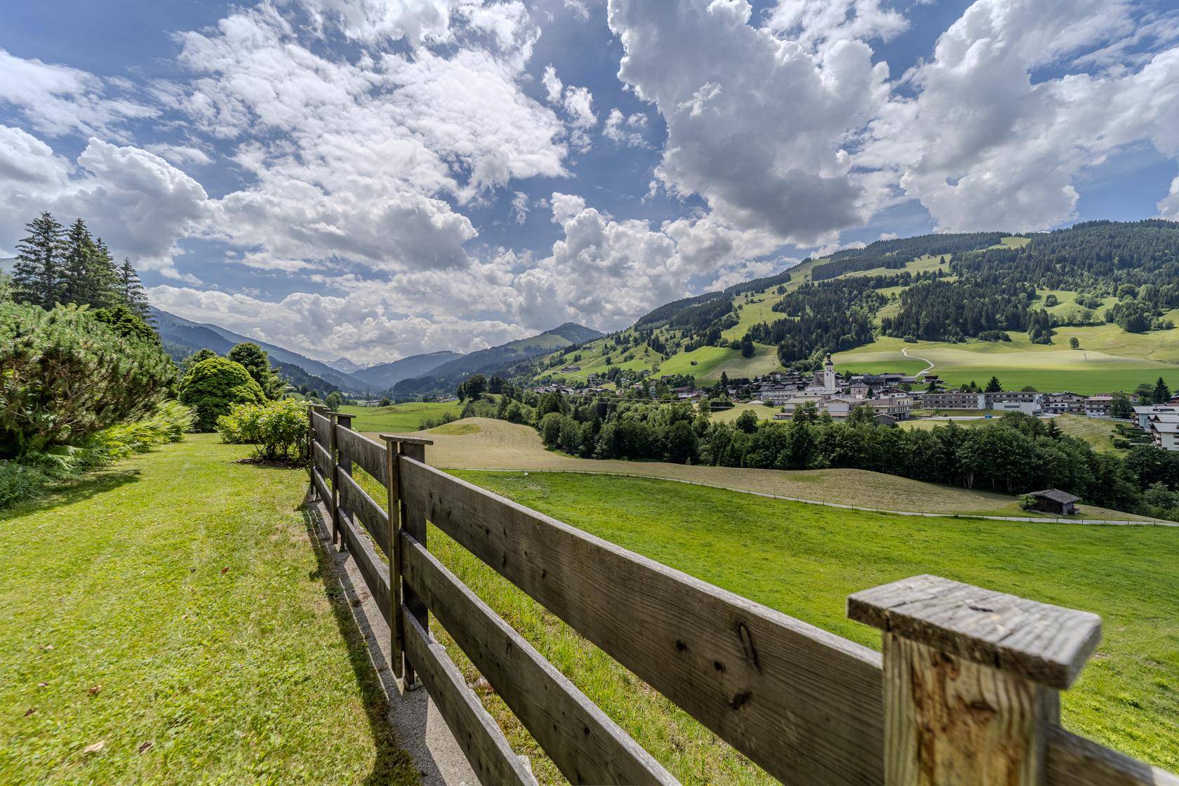 Malerische Landschaft mit Holzzaun, grünen Wiesen und einem Dorf in der Ferne unter blauem Himmel.