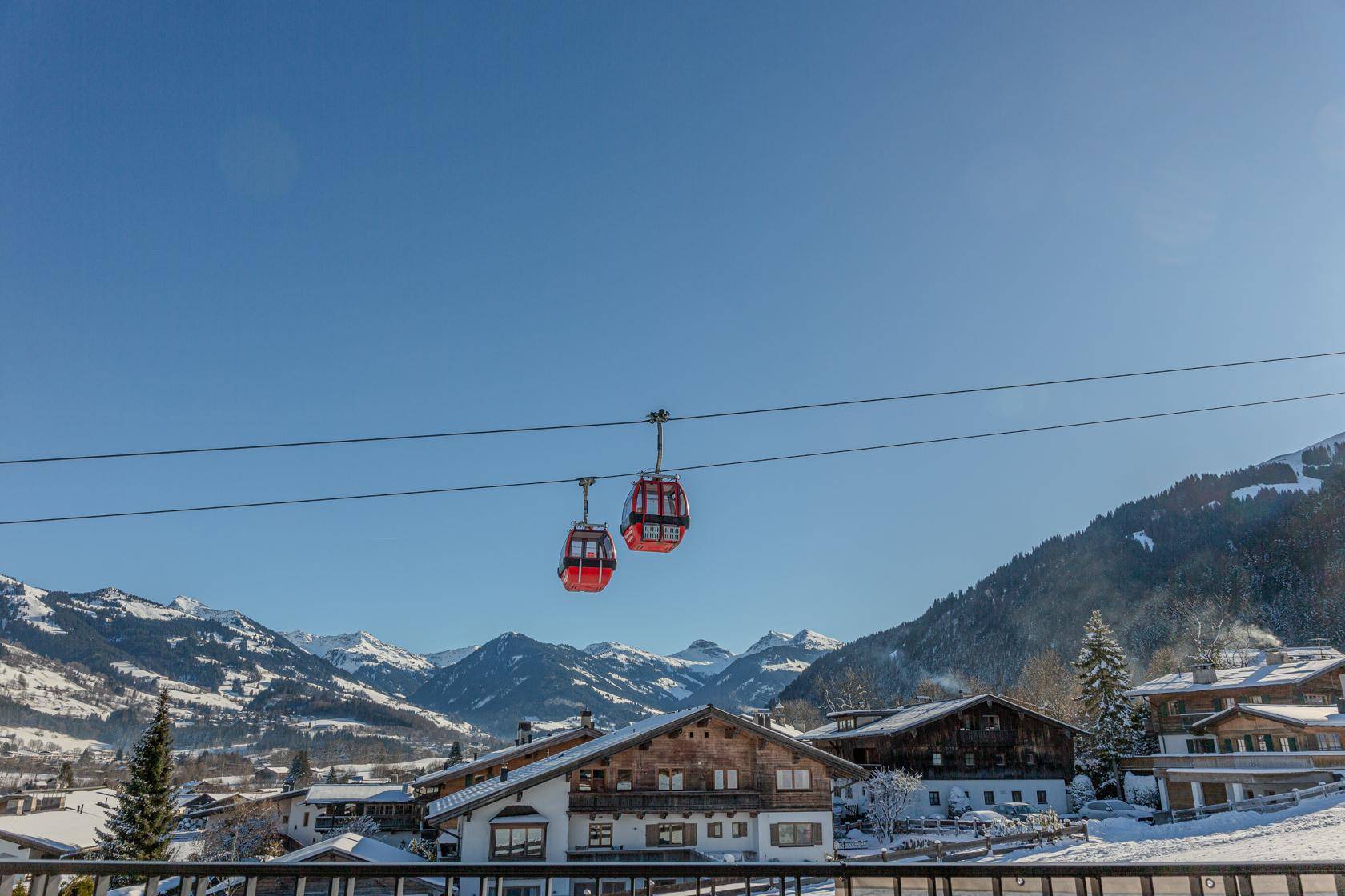 Panoramablick auf verschneite Berge und traditionelle Häuser mit Skiliftkabinen im Vordergrund.