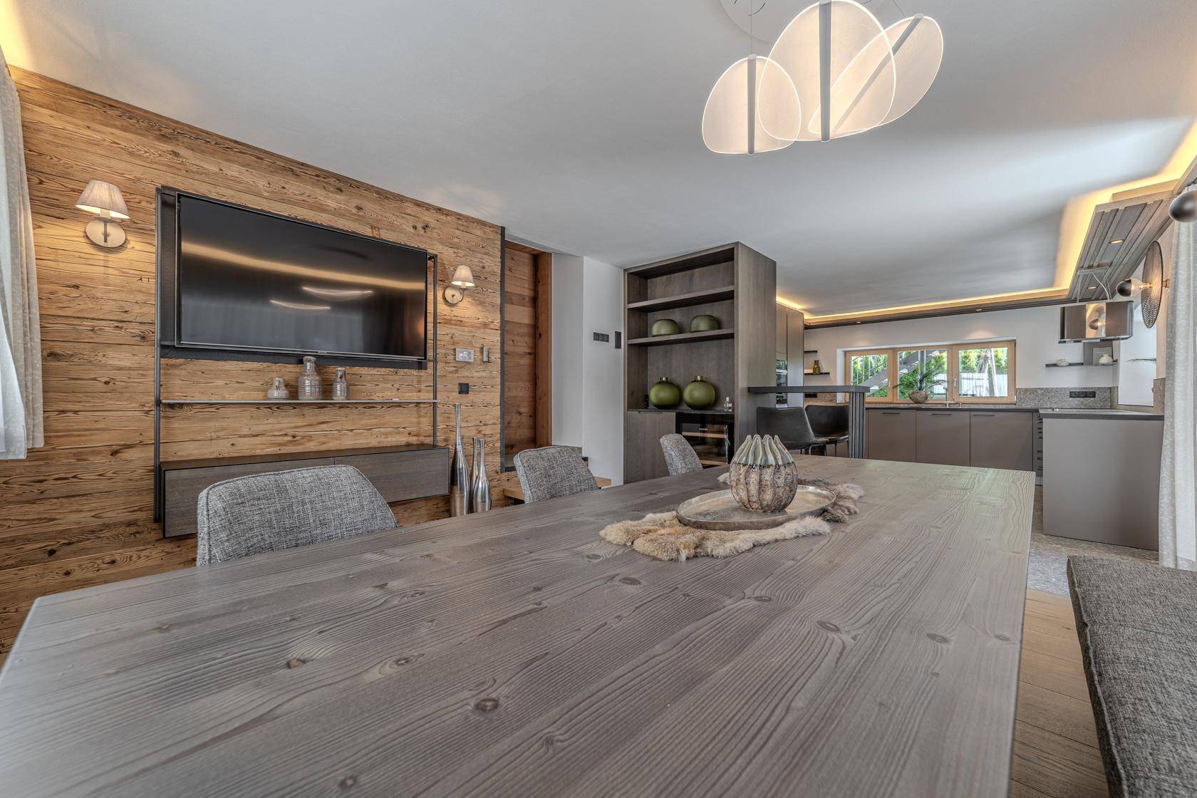 Offener Wohn- und Essbereich mit Holzwand, Fernseher und moderner Küche im Hintergrund.