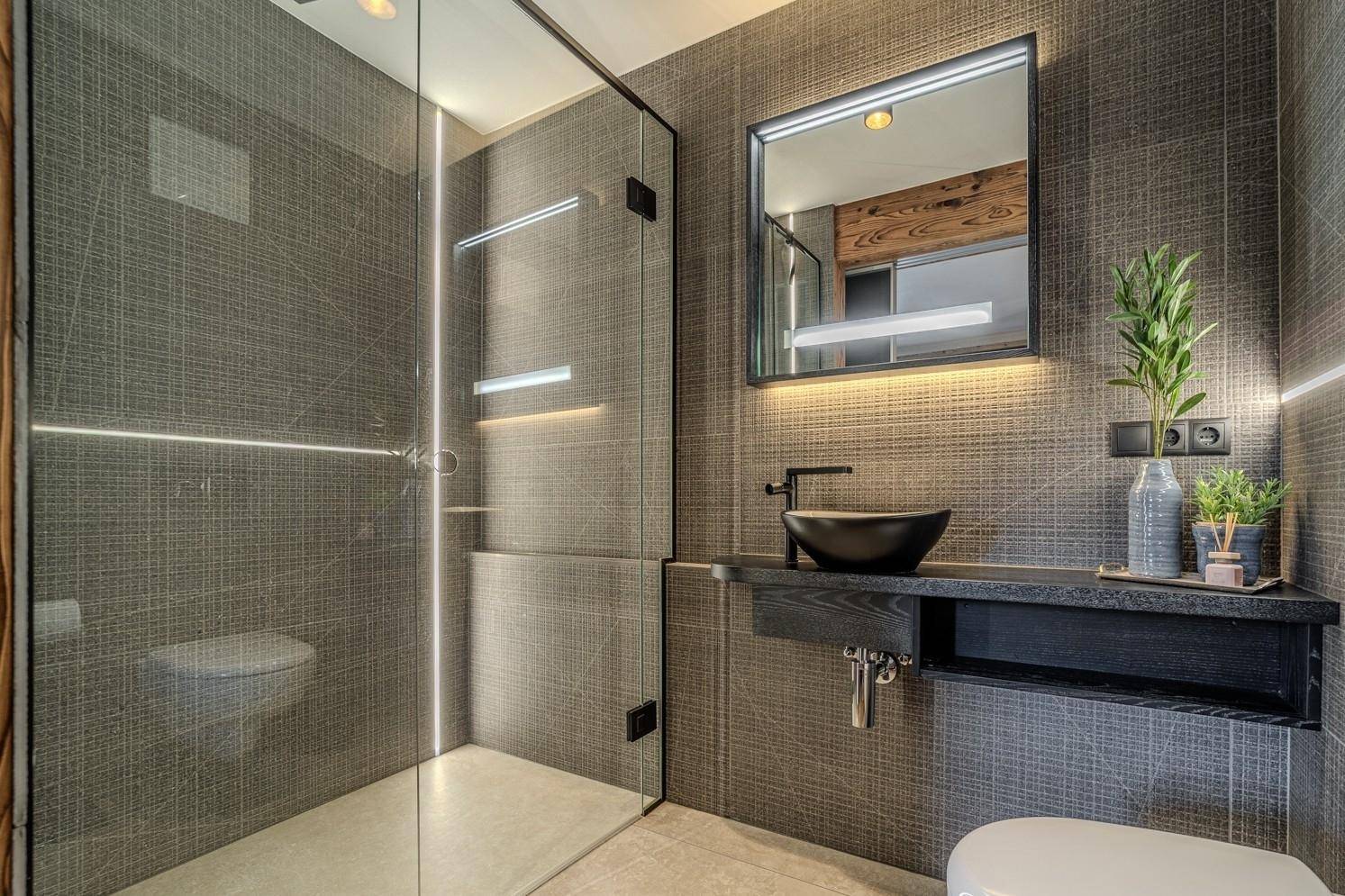 Elegantes Badezimmer mit dunklem Waschbecken, beleuchtetem Spiegel und geräumiger Dusche.