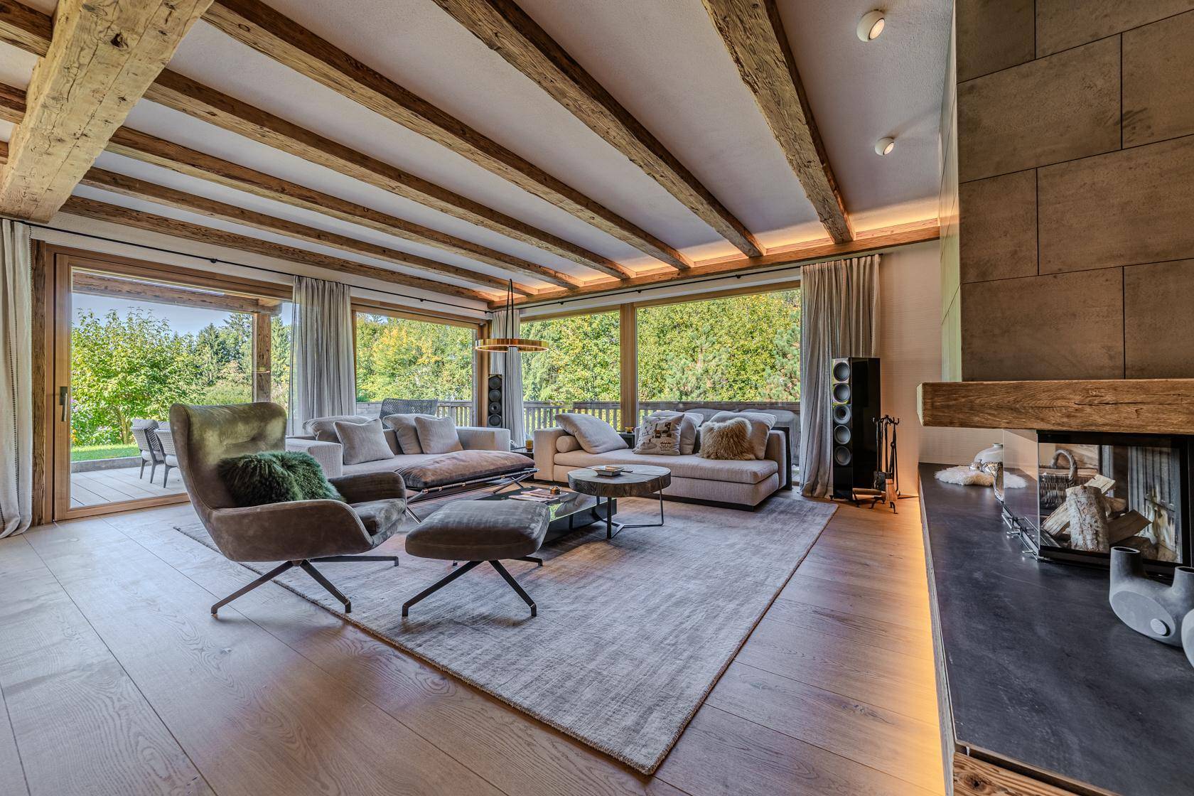 Helles Wohnzimmer mit Holzbalkendecke, Kamin und großen Fenstern mit Blick ins Grüne.