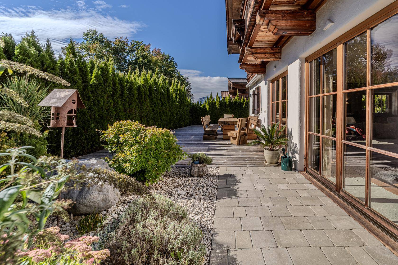 Gepflegter Gartenbereich mit Steinbeet, Hecke und Vogelhaus, angrenzend an eine gepflasterte Terrasse.